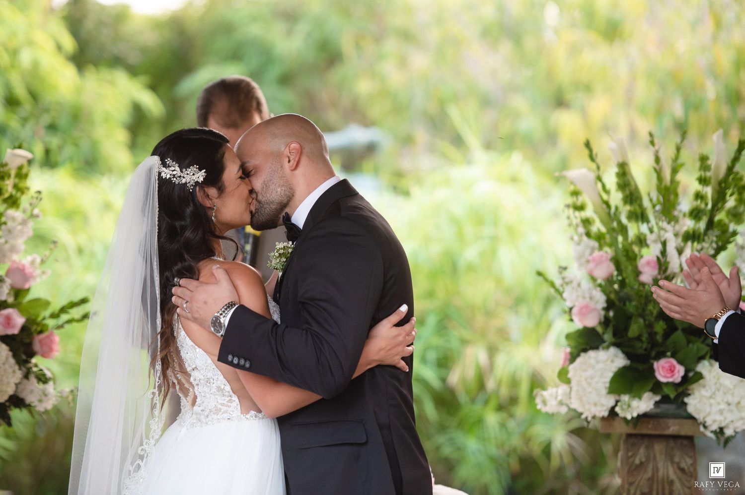 Bride and groom share their first kiss at an outdoor wedding ceremony surrounded by lush greenery and pink floral arrangements.