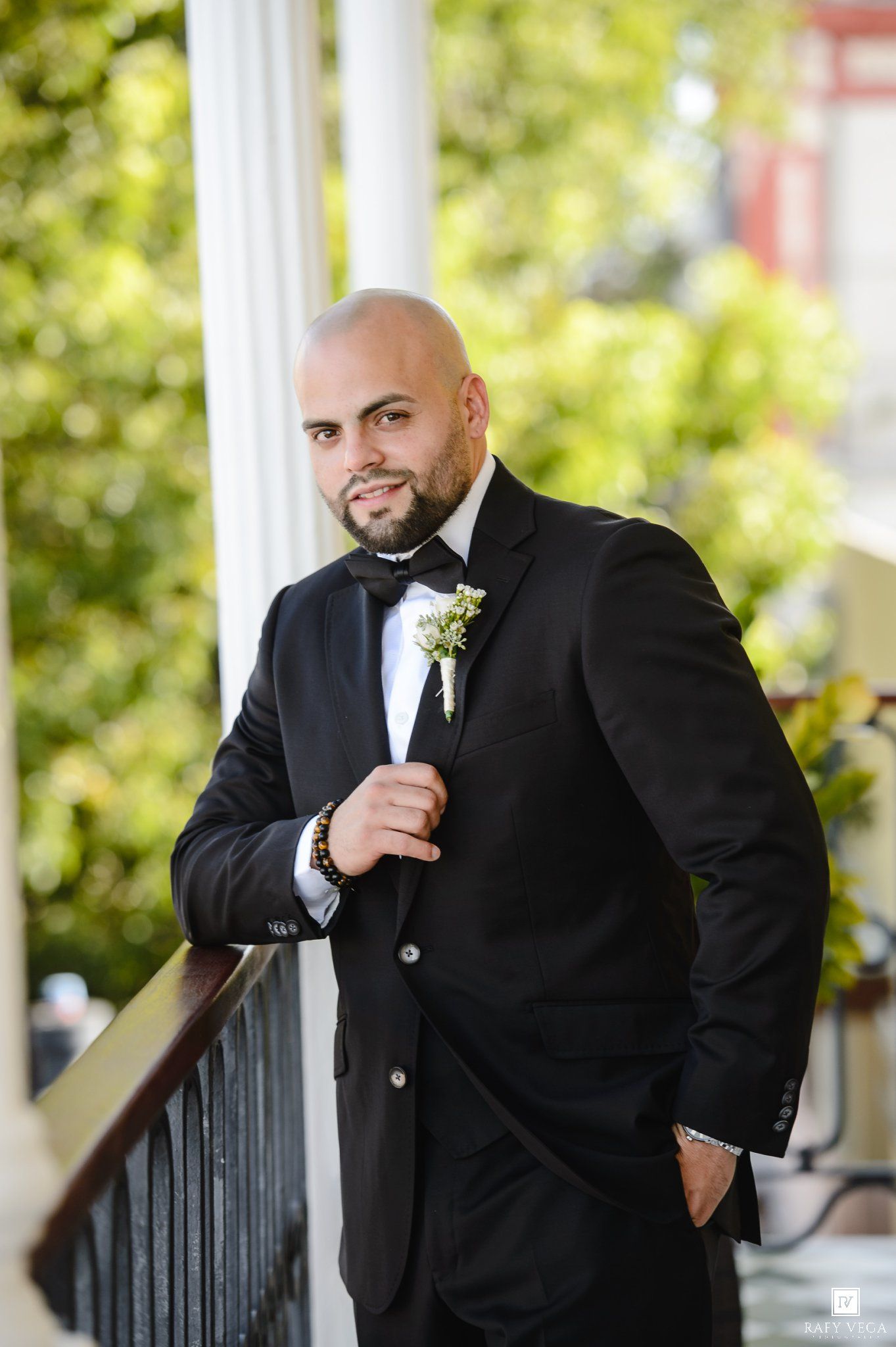 Bald groom in black tuxedo with boutonniere poses on elegant white-columned balcony surrounded by lush greenery.