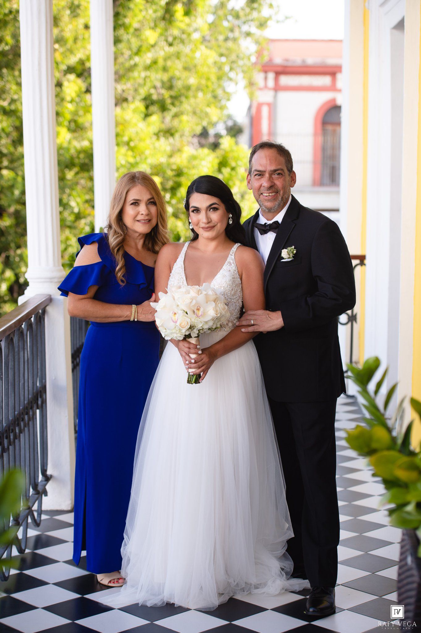 Bride in white gown holding bouquet poses with parents on checkered balcony at elegant wedding venue.