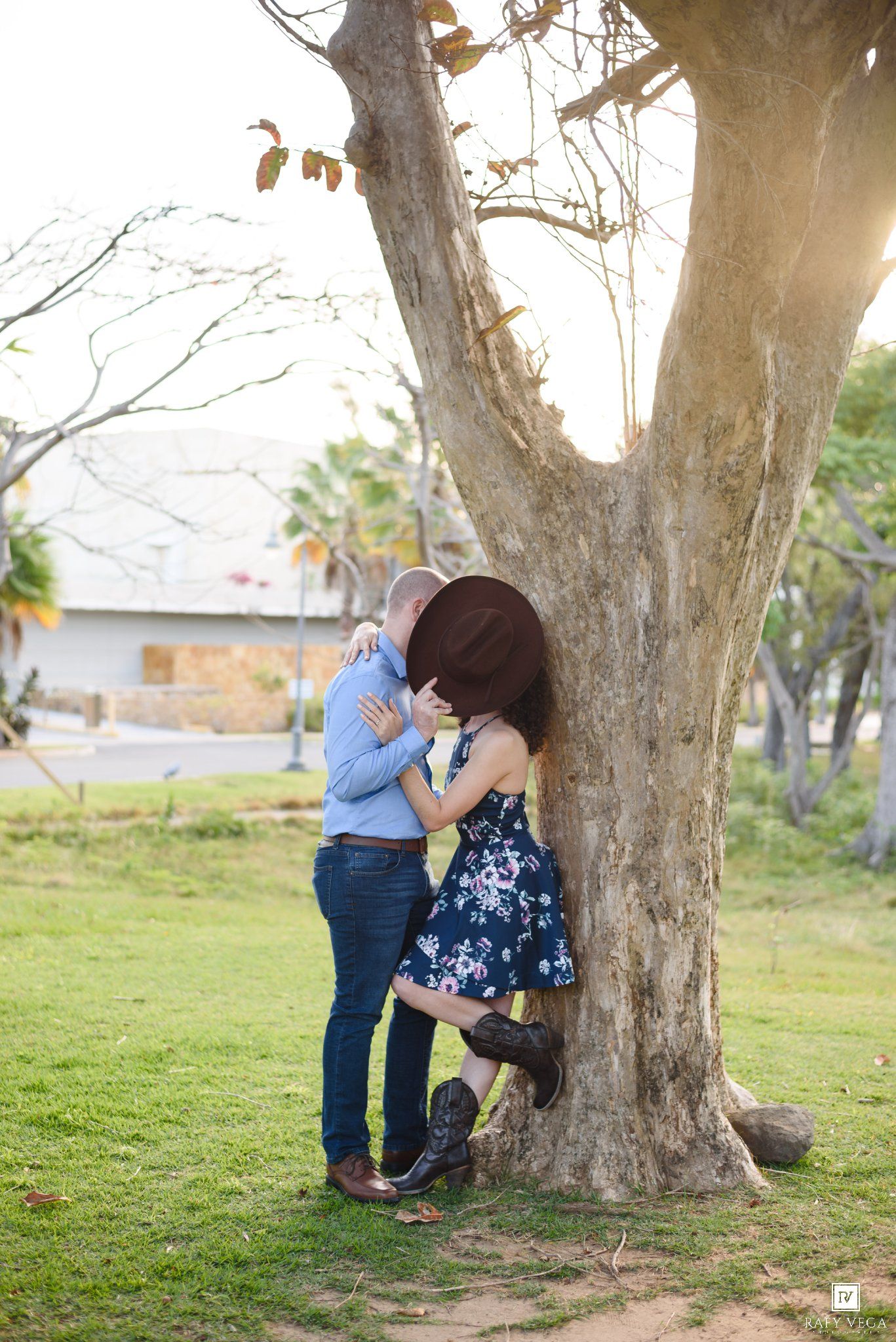 Couple sharing a romantic kiss leaning against a large tree in a sunny park setting.