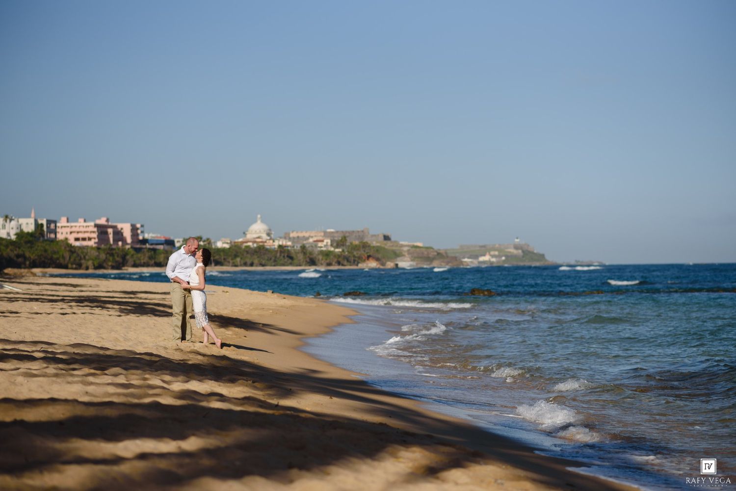 Couple embracing on a sandy beach with turquoise ocean waves and a scenic coastal town in the background.