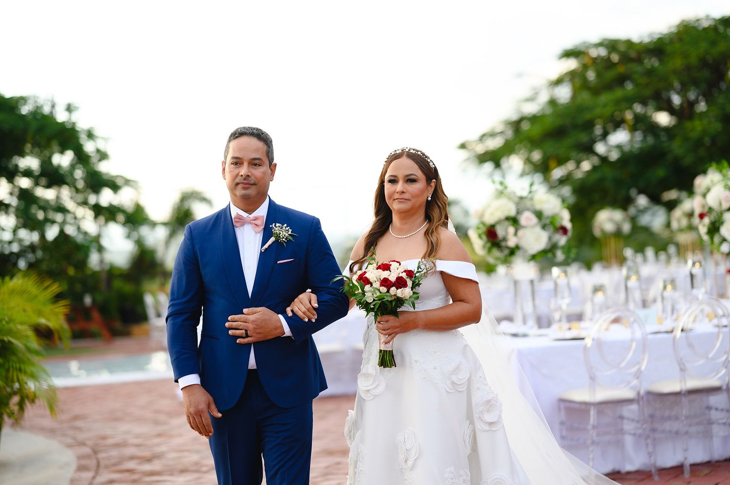 Bride in white off-shoulder gown holding red bouquet walks with groom in blue suit at outdoor wedding reception.