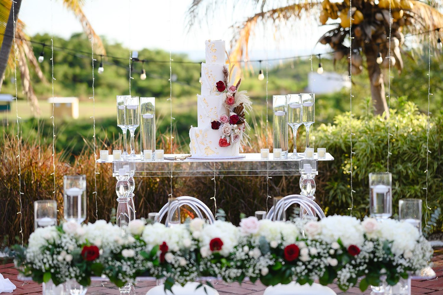 Elegant outdoor wedding dessert table with white tiered cake, champagne flutes, and red and white floral arrangements.