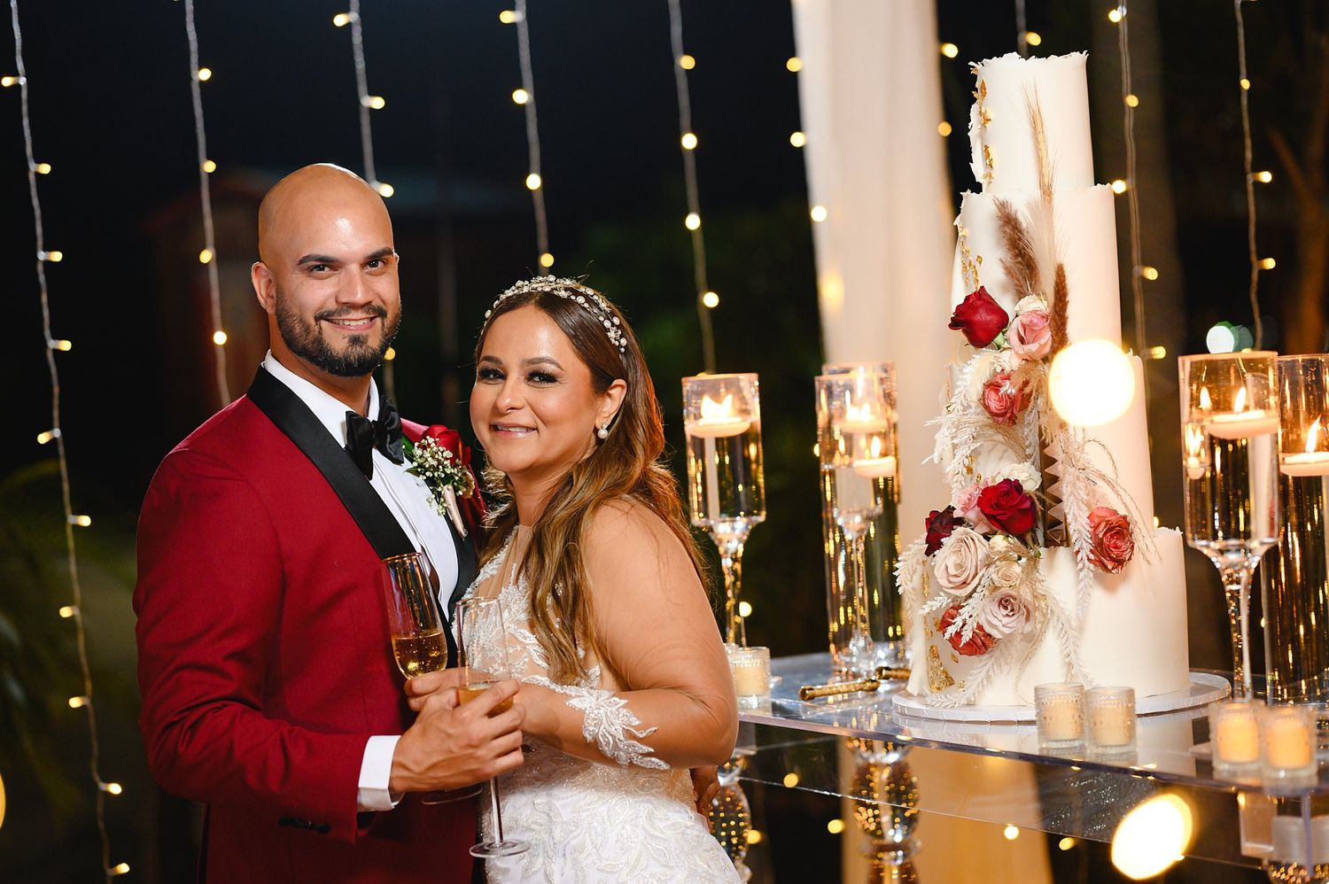 Bride and groom smiling together beside elegant white wedding cake with floral decor and fairy lights backdrop.