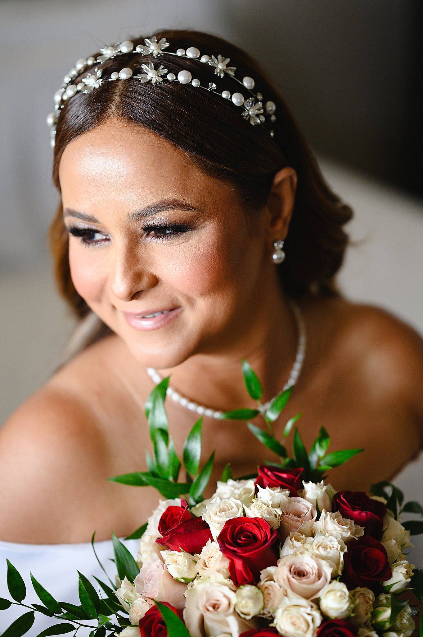 Bride smiling with pearl headpiece, diamond earrings, and bouquet of red and white roses on her wedding day.
