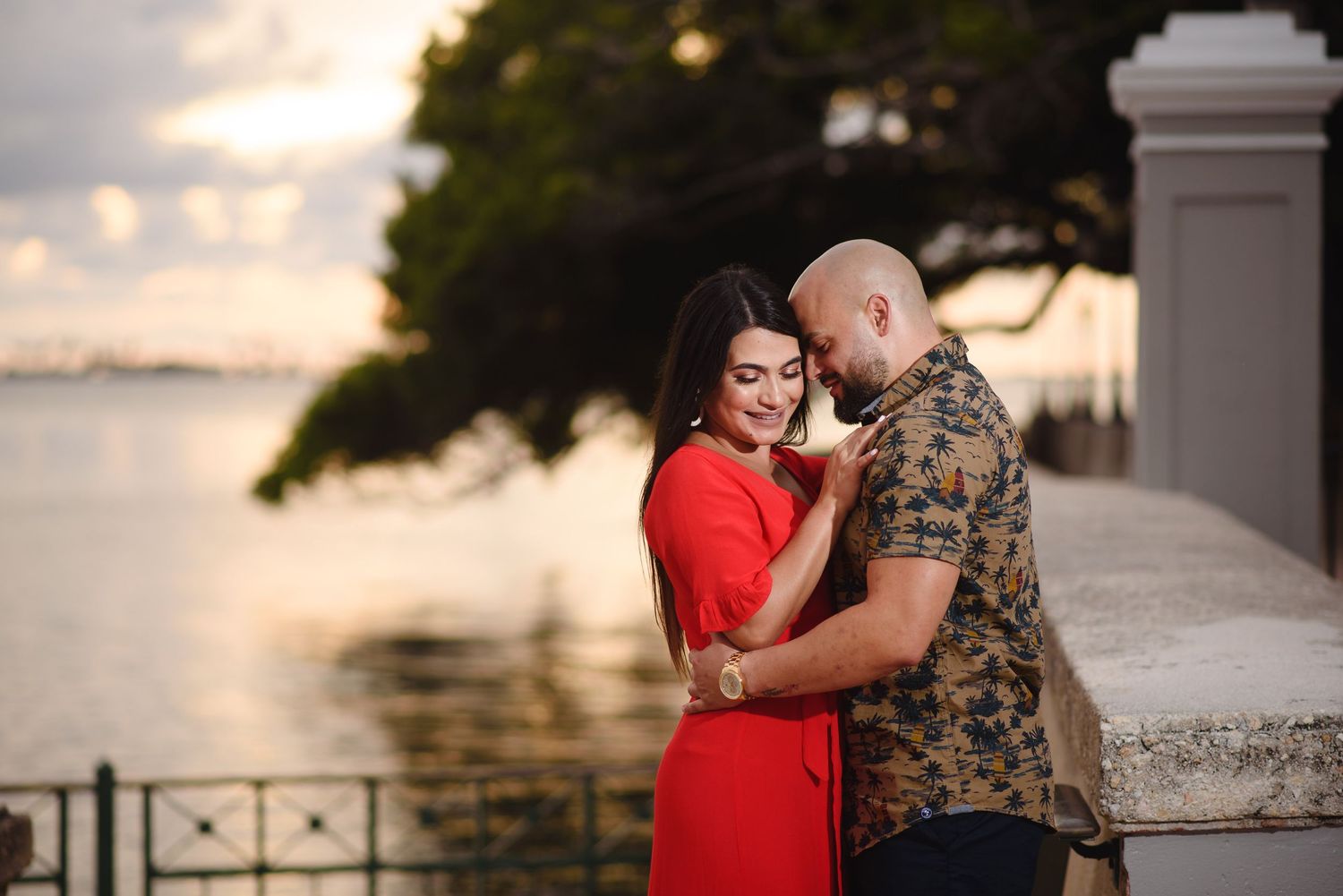 Couple sharing a romantic embrace at sunset by the waterfront, woman in red dress and man in floral shirt.
