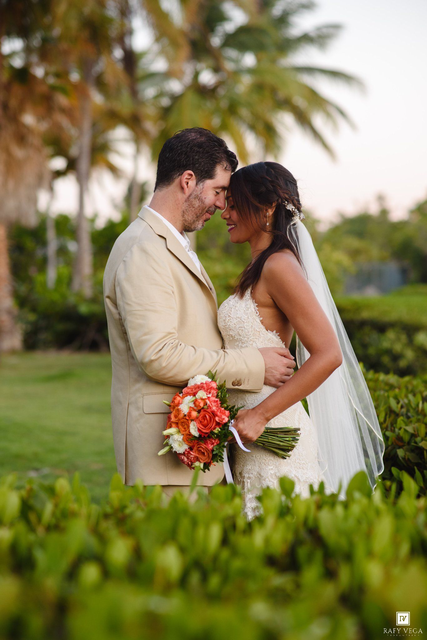 Smiling bride and groom share romantic moment face-to-face in lush tropical garden at sunset.