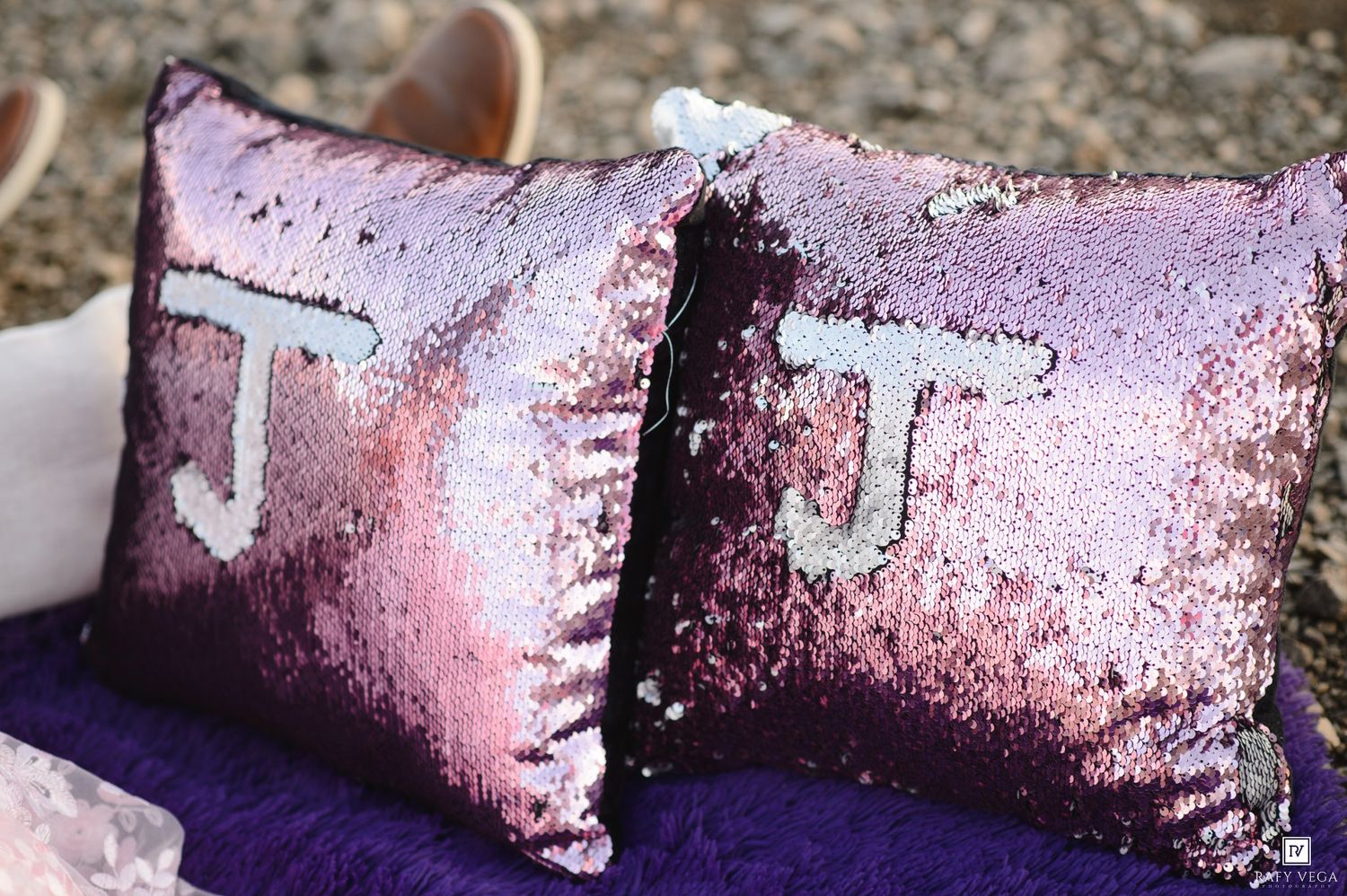 Two pink sequin throw pillows with letter J monograms displayed on a purple fabric surface outdoors.