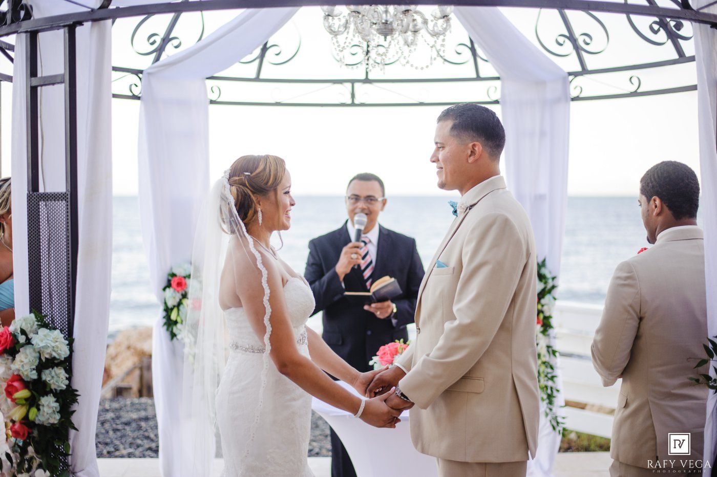 Couple exchanges vows at outdoor beach wedding ceremony under white arch with officiant and ocean backdrop.