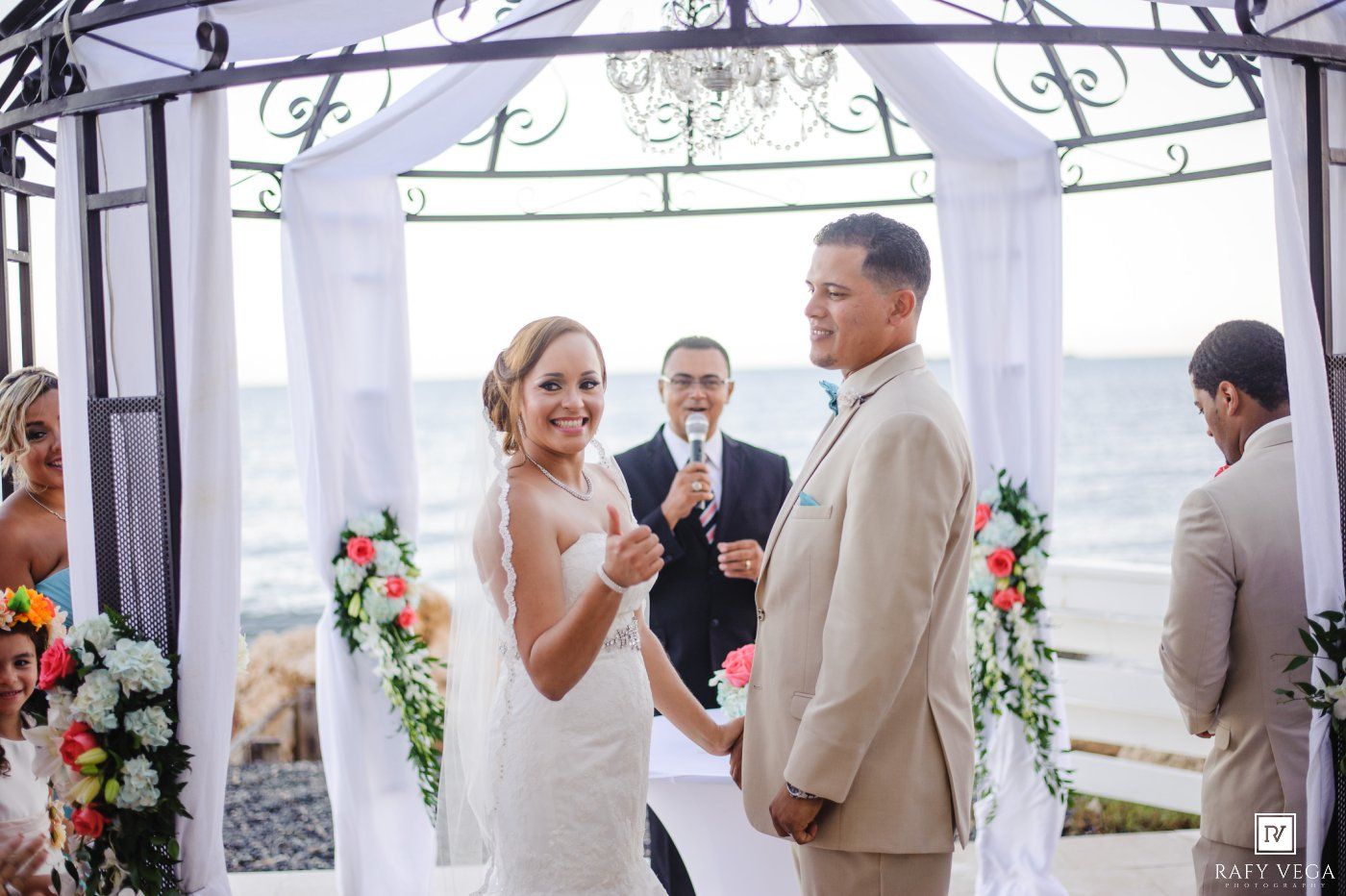 Smiling bride exchanges vows with groom at beachside gazebo ceremony with officiant and colorful floral decorations.