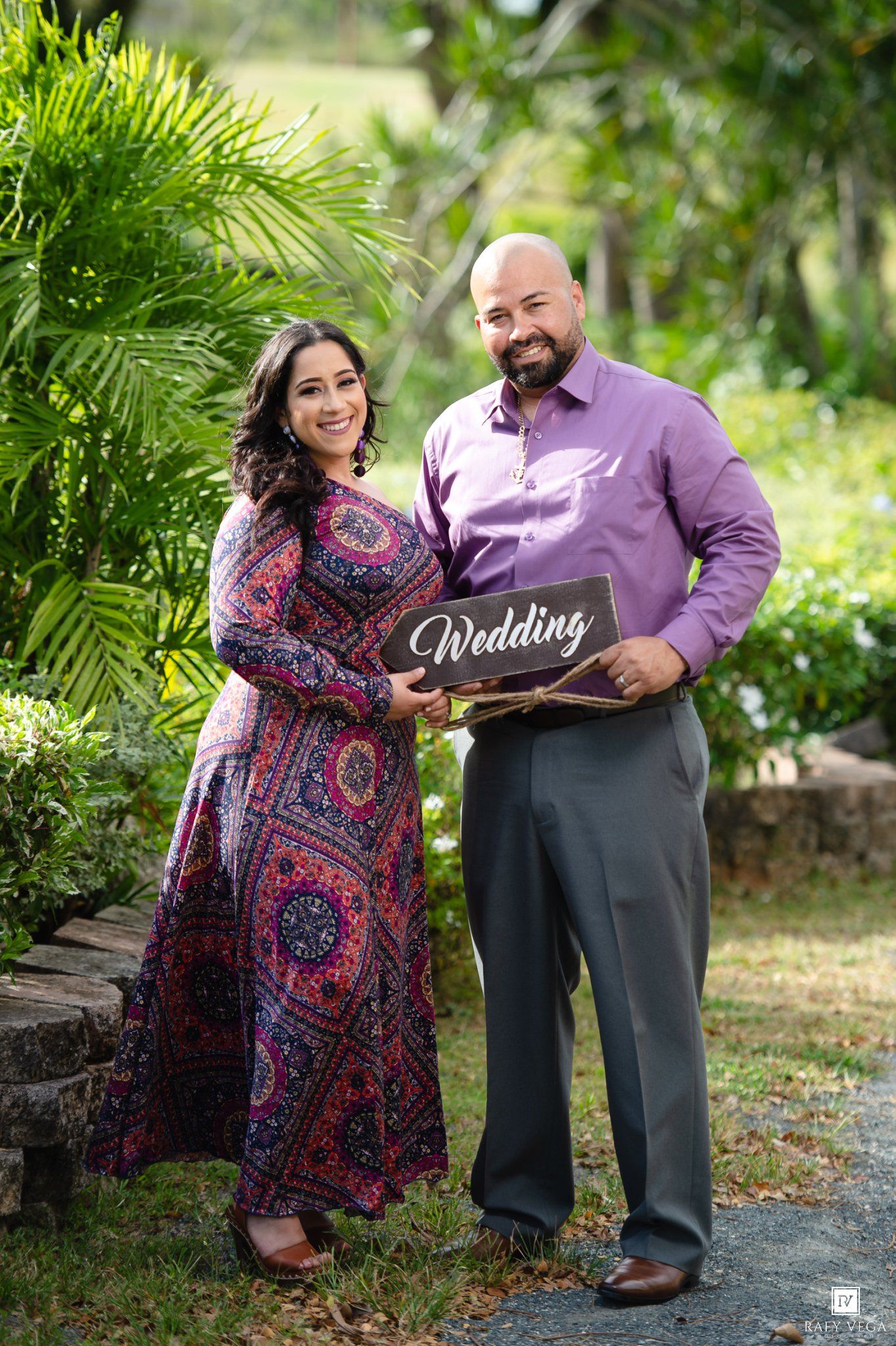 Couple smiling and holding a Wedding sign outdoors, woman in floral dress and man in purple shirt and gray pants.