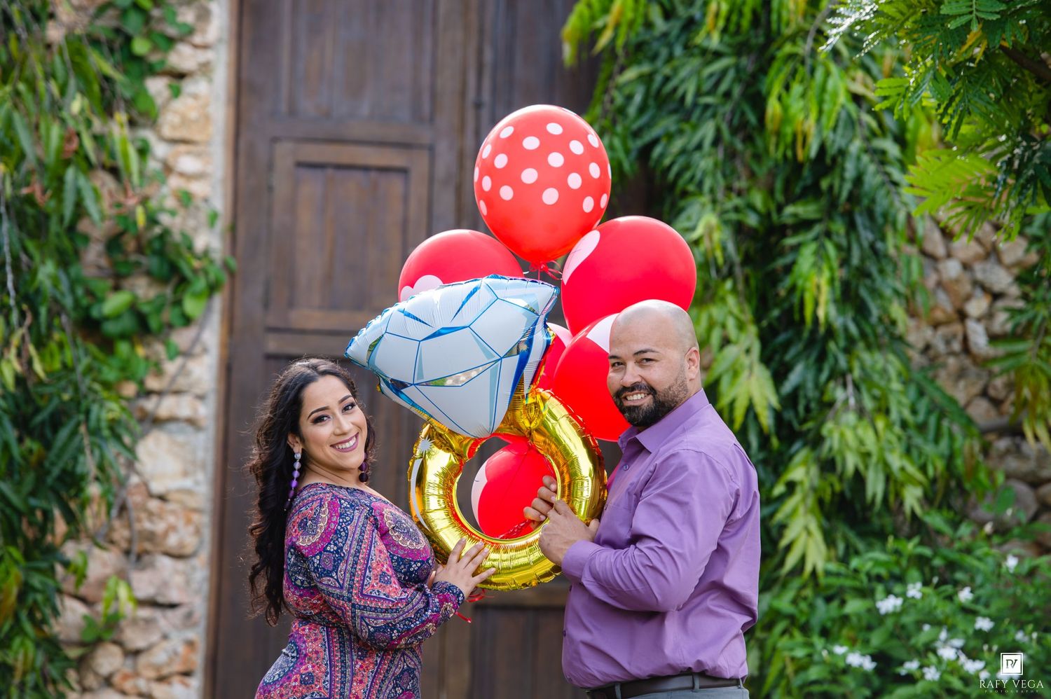 Couple smiling outdoors holding colorful balloons including red polka dot, gold number, and diamond ring shapes.