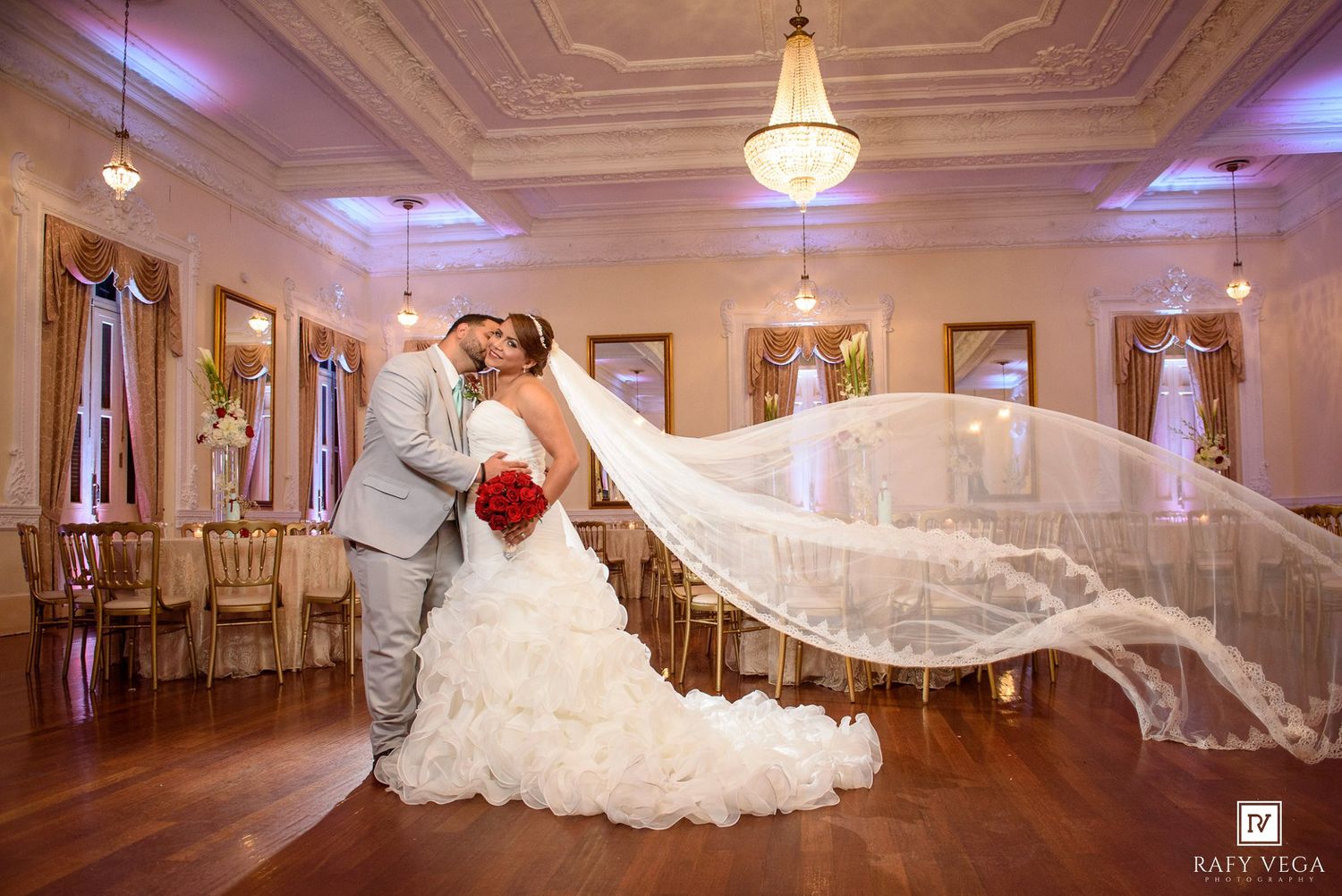 Bride and groom share a romantic kiss in an elegant ballroom, with her flowing cathedral veil dramatically billowing behind her.