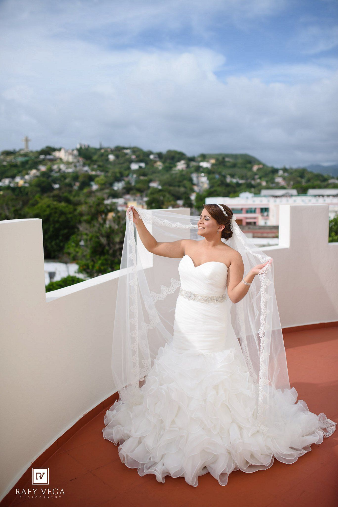 Bride in mermaid wedding gown holding veil on rooftop with scenic hillside view.