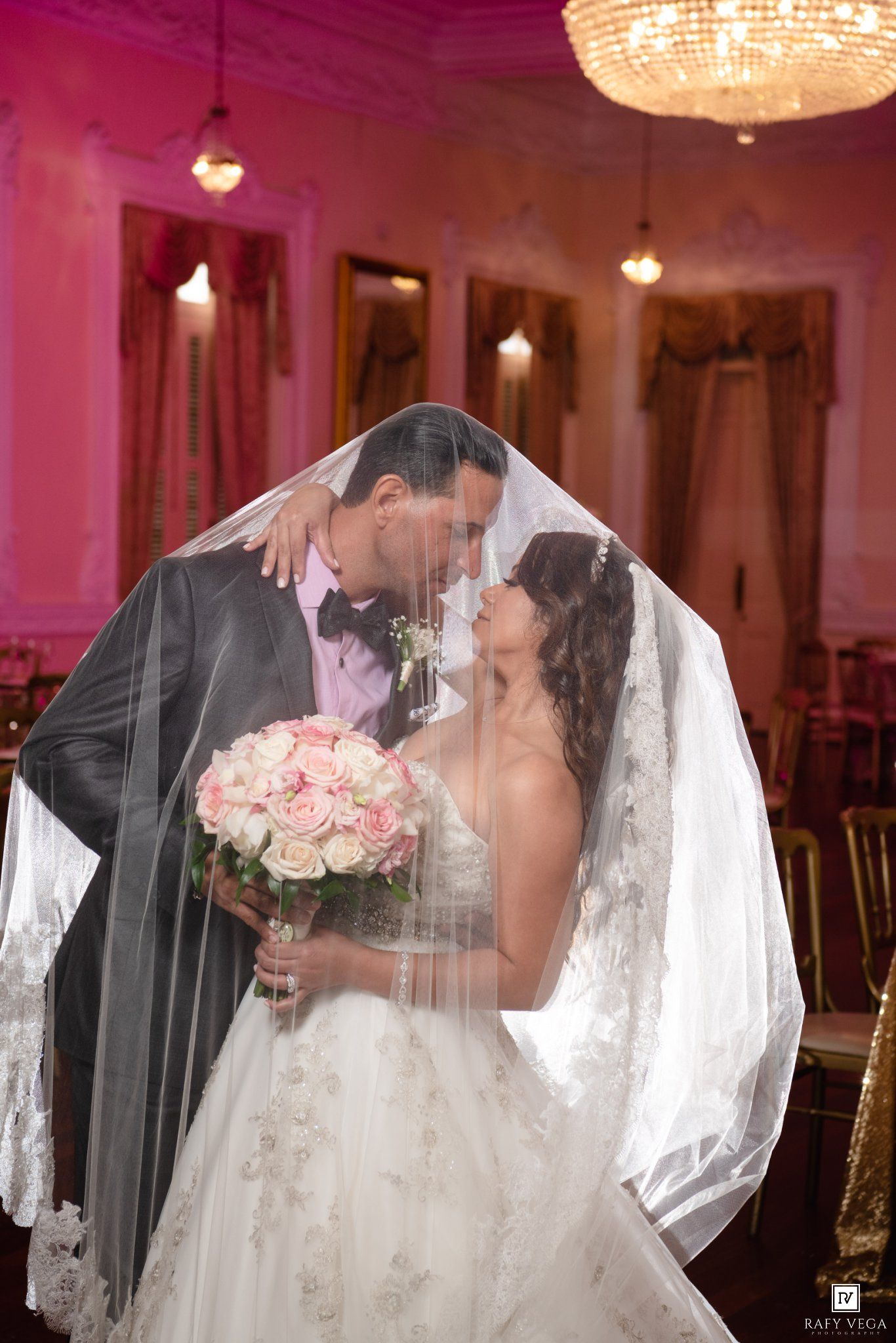 Couple shares a kiss under the bride's veil, bride holding a pink rose bouquet in a grand ballroom.