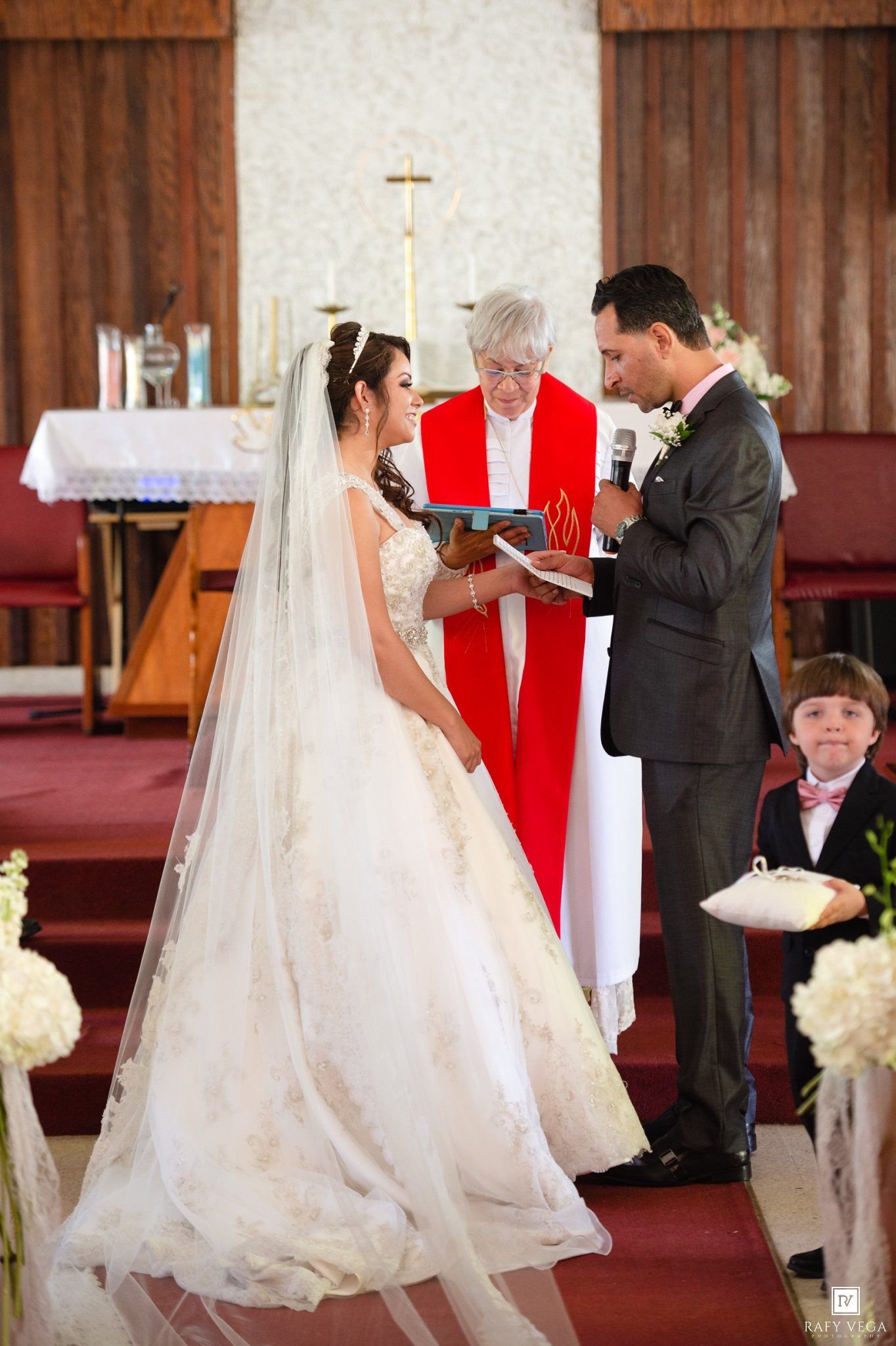 Bride and groom exchange vows at church altar with officiant in red stole and ring bearer nearby.