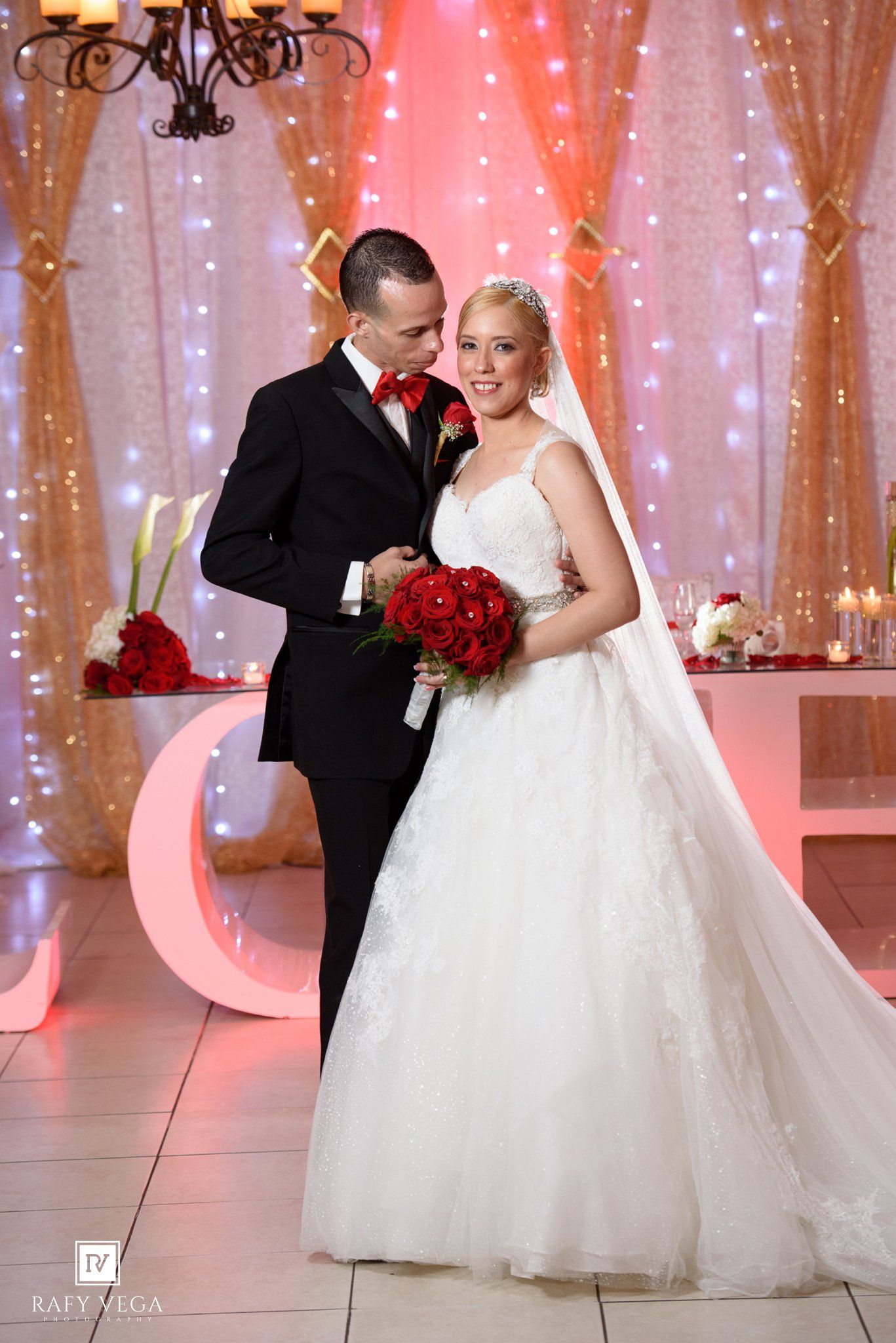 Bride in white ballgown holding red roses bouquet stands with groom in black tuxedo and red bow tie at elegant wedding venue.