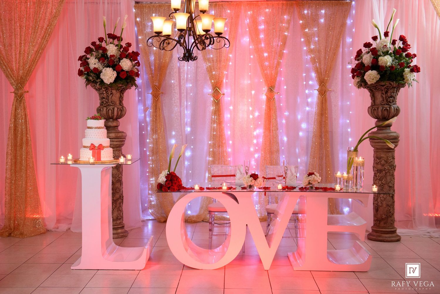 Elegant wedding reception backdrop with LOVE letters, floral arrangements, chandelier, fairy lights, and a white cake display.