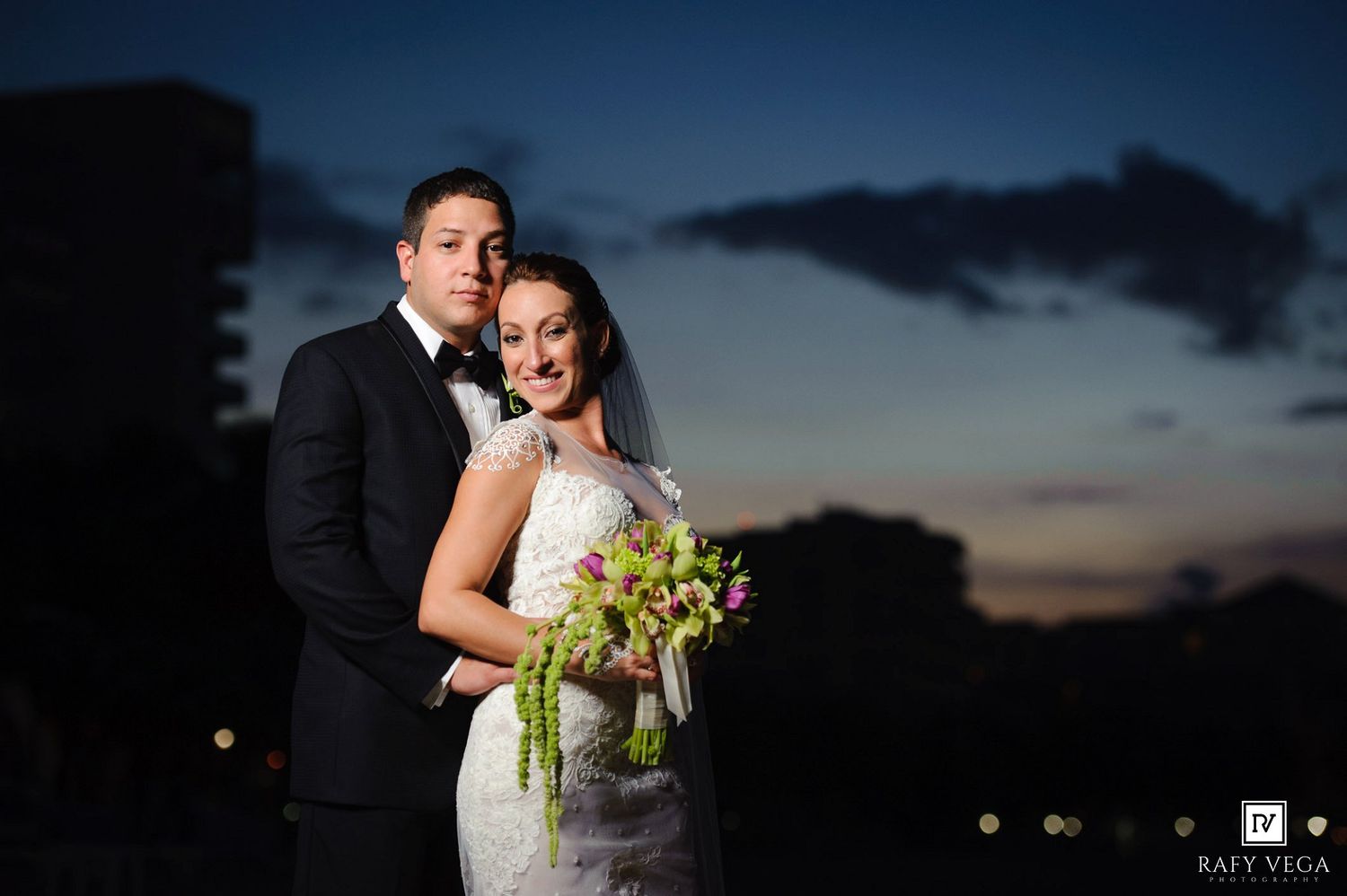 Bride and groom pose together at twilight, with the bride holding a green floral bouquet in a lace wedding gown.
