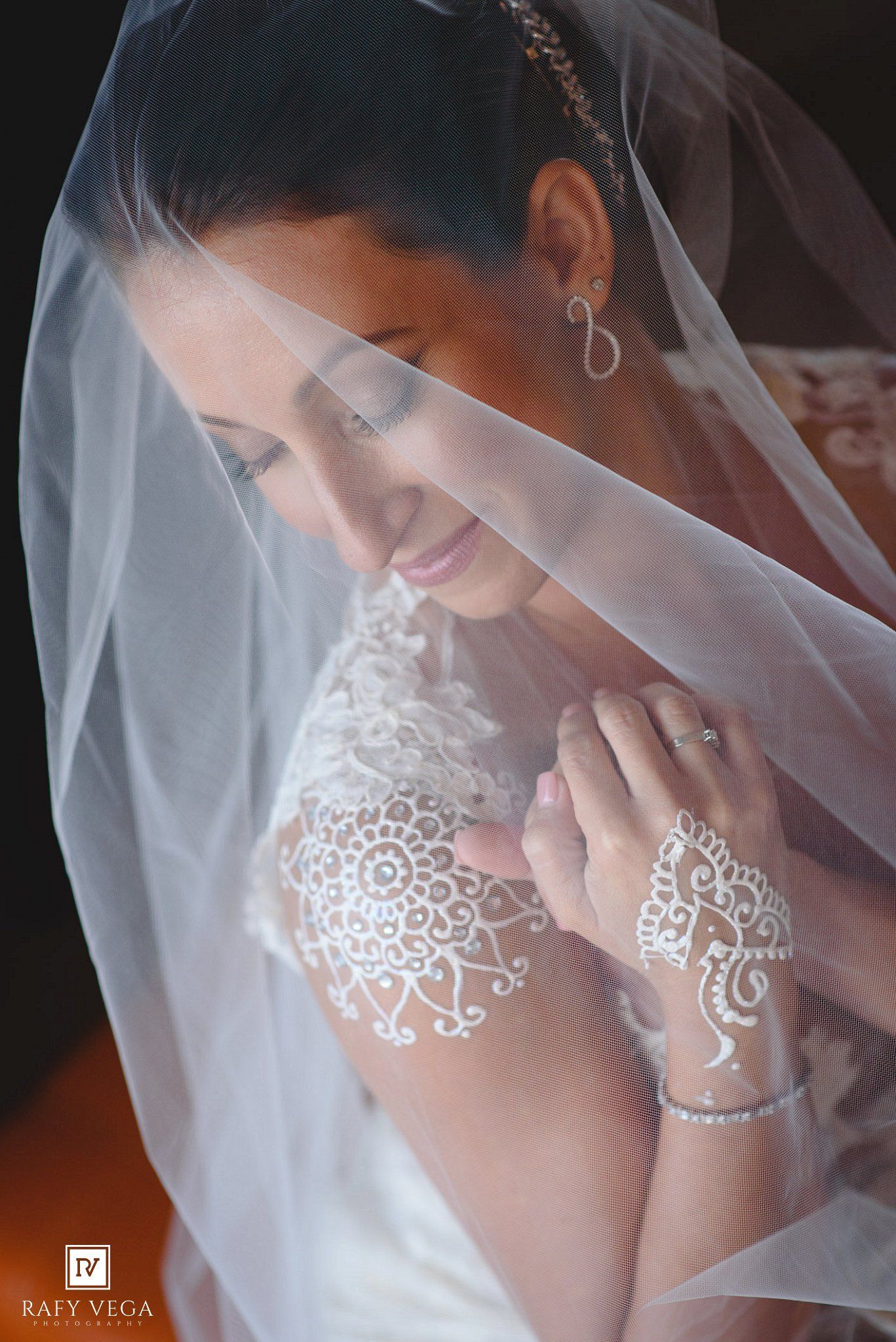 Bride in white lace gown and veil with elegant white henna on hand, smiling softly on her wedding day.