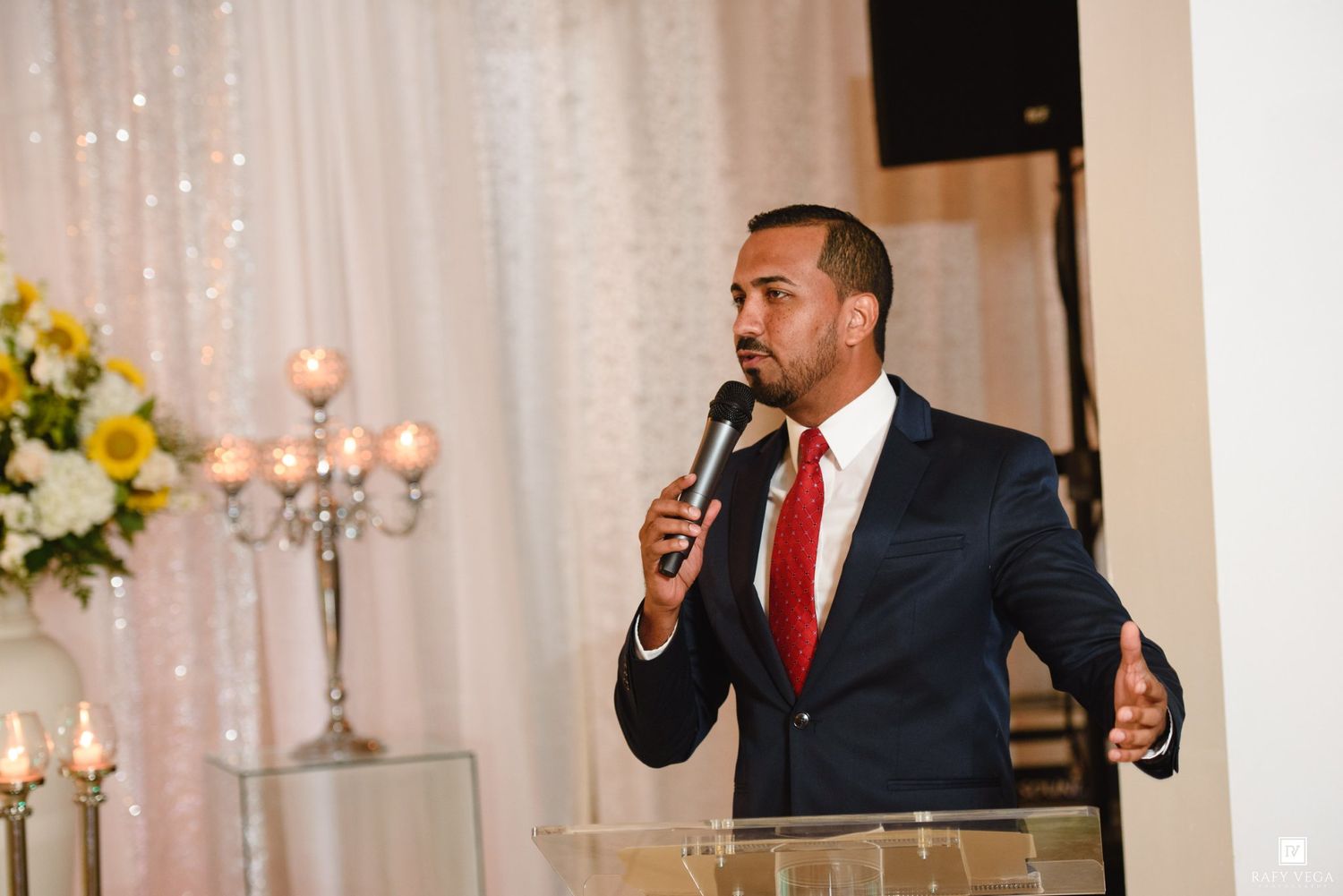 Man in navy suit with red tie speaking into microphone at elegant wedding reception with floral backdrop.