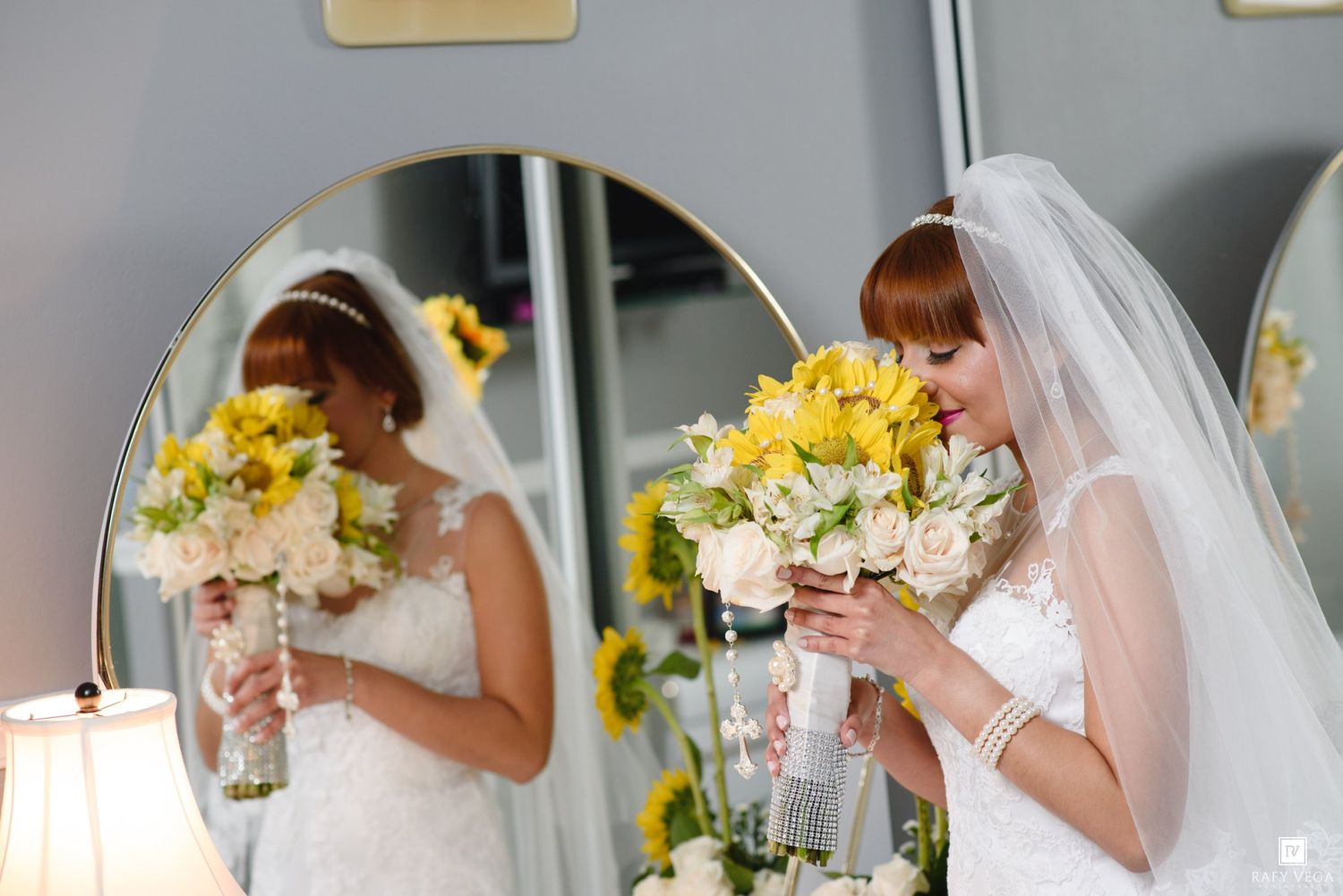 Bride in white gown holds yellow sunflower bouquet reflected in round mirror.