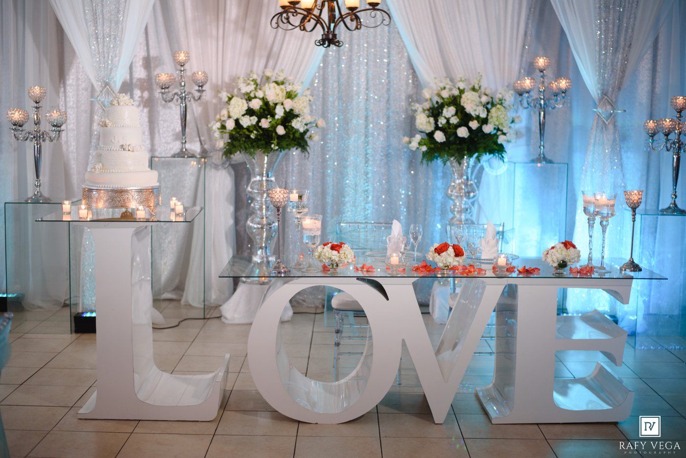 Elegant wedding reception dessert table with large LOVE letters, white cake, floral arrangements, and candles in a draped venue.
