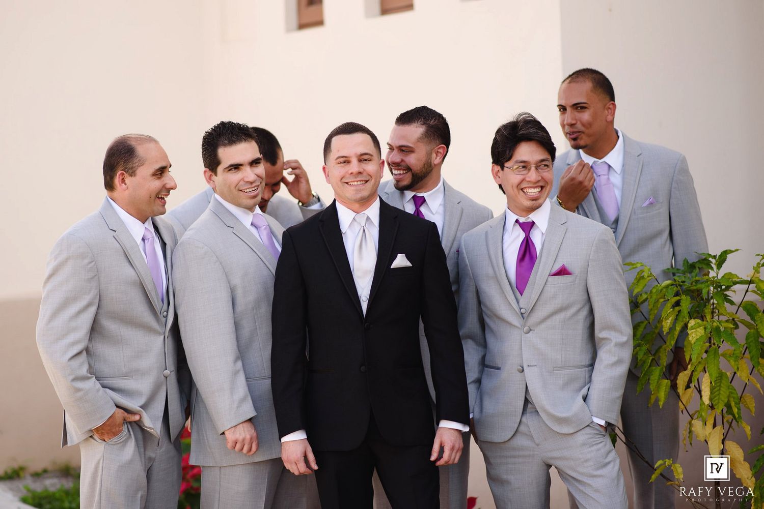 Groom in black tuxedo poses with groomsmen wearing gray suits and purple ties outside wedding venue.