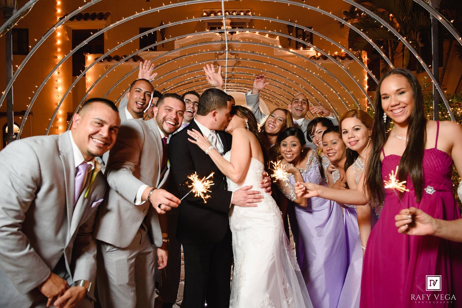 Wedding party celebrates with sparklers under a lit arch as bride and groom share a romantic kiss.