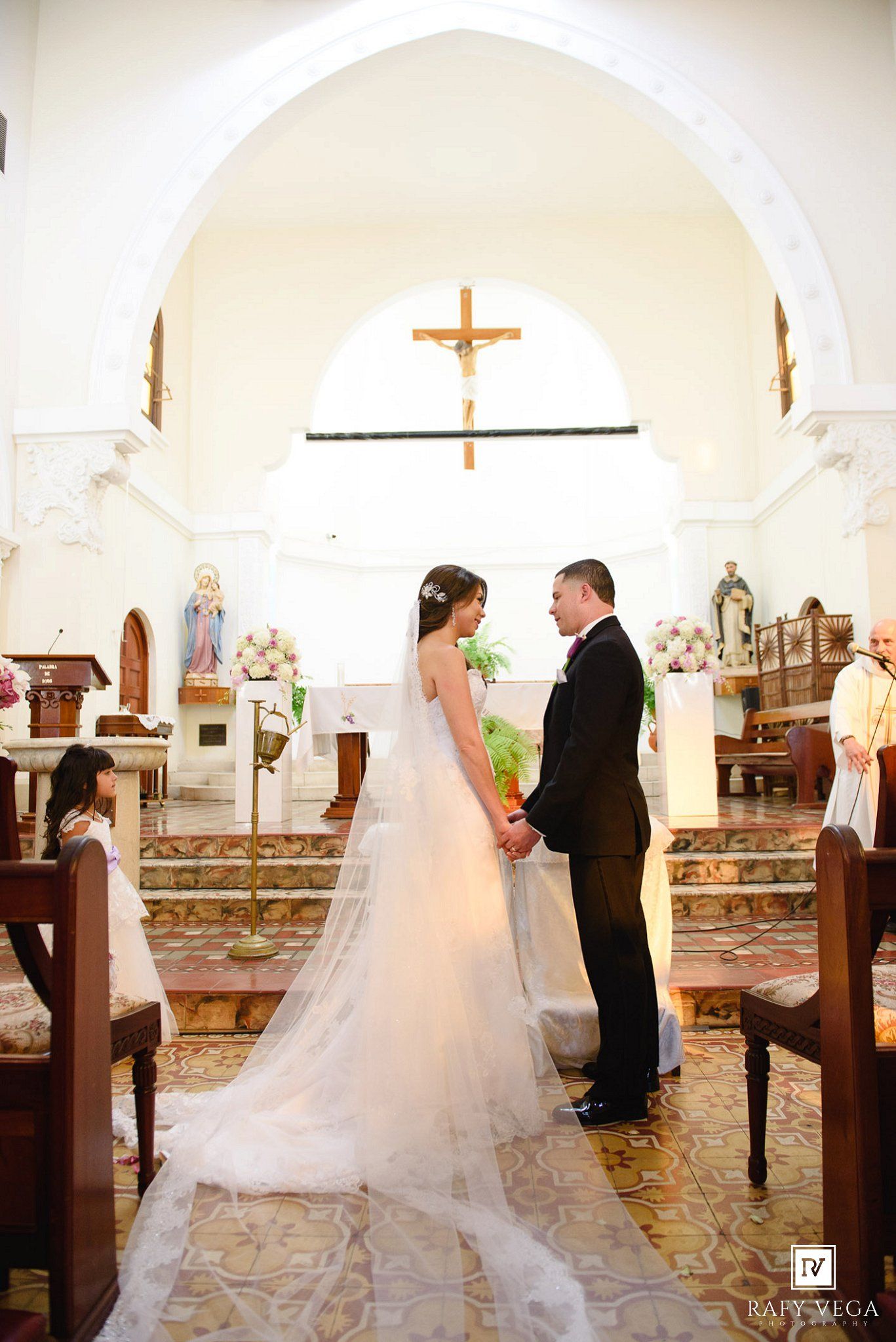 Bride and groom hold hands at the altar during a Catholic church wedding ceremony with a crucifix backdrop.