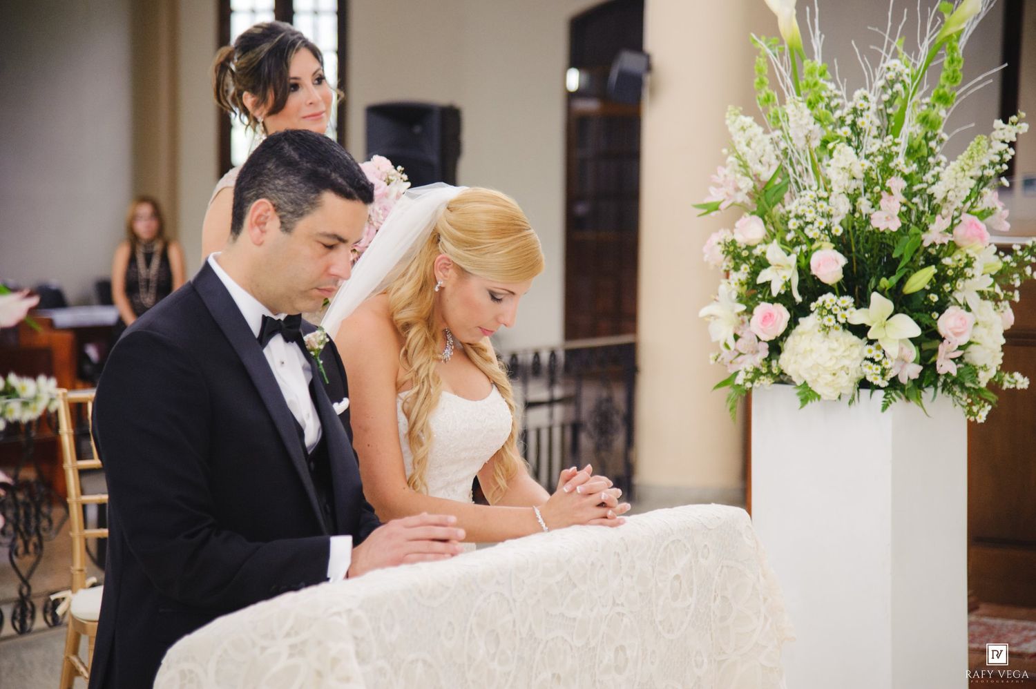 Bride and groom kneeling in prayer at elegant church wedding ceremony with white floral arrangements.