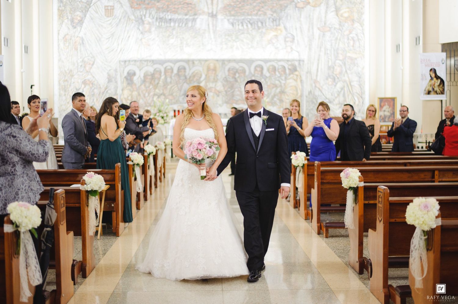 Bride and groom walk down church aisle after wedding ceremony, surrounded by guests and floral decorations.