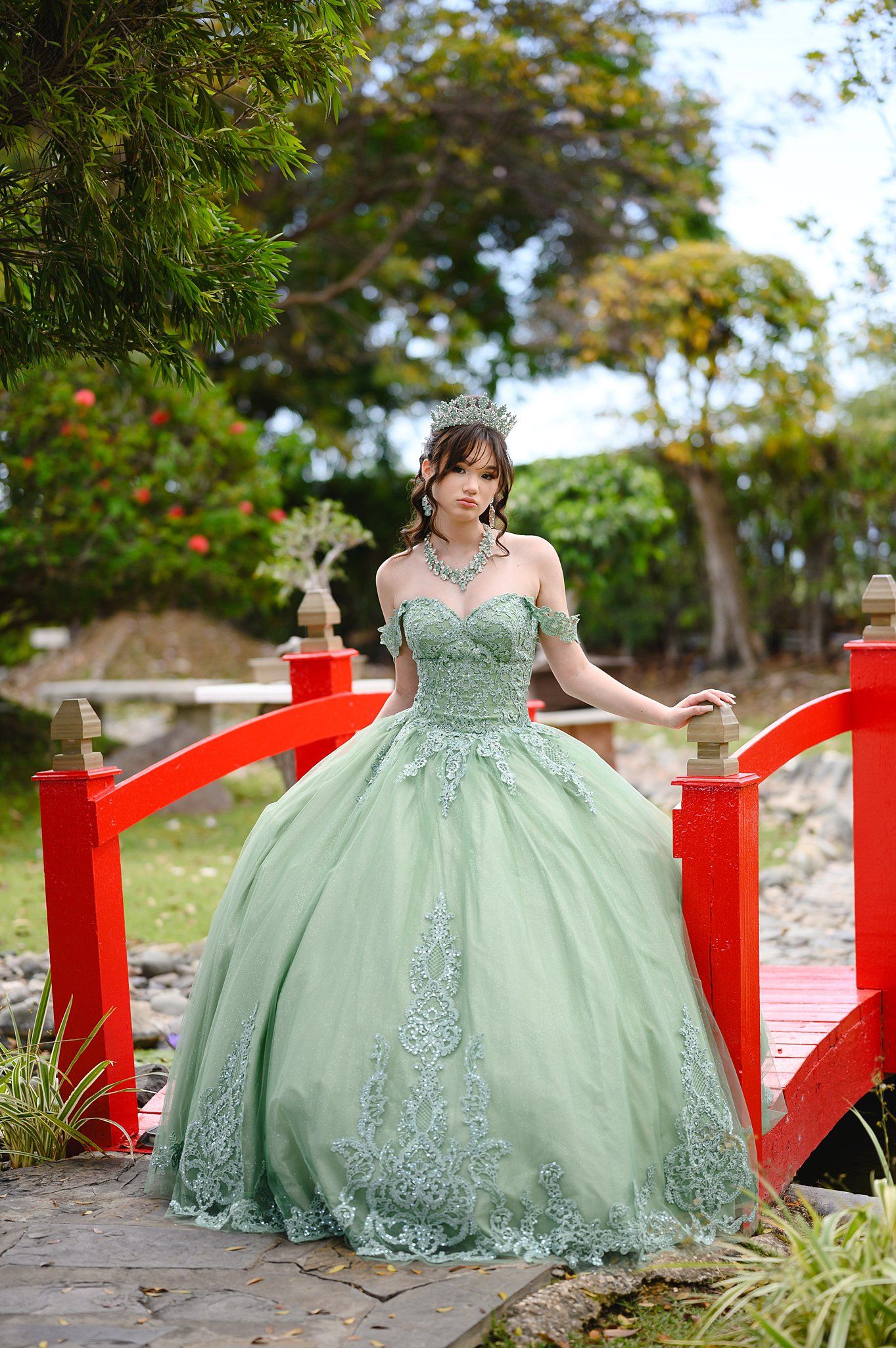 Young woman in elegant sage green ball gown with lace details and tiara poses on red bridge in lush garden.