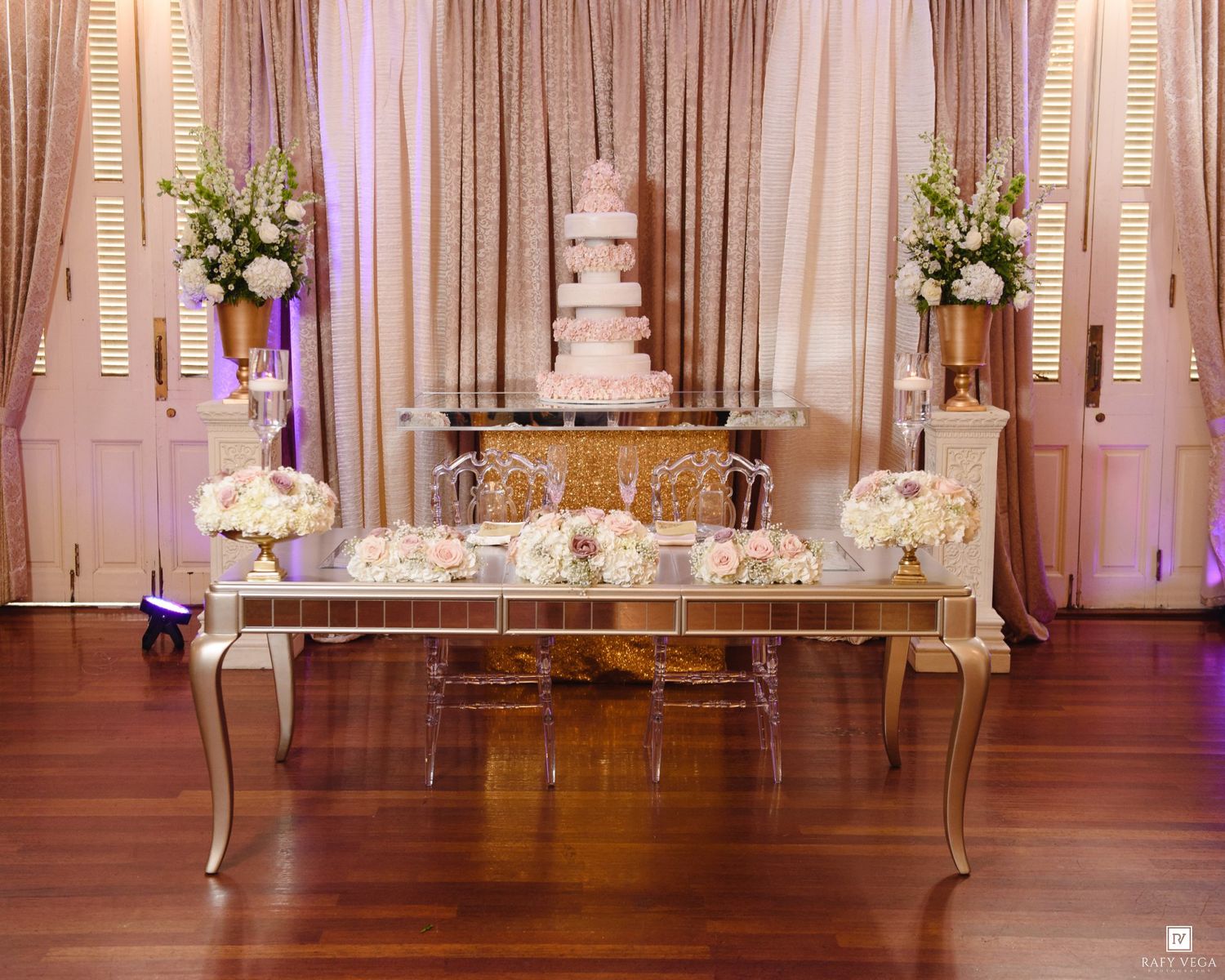 Elegant wedding cake table with mirrored display, floral arrangements, and a tiered cake against draped curtains.