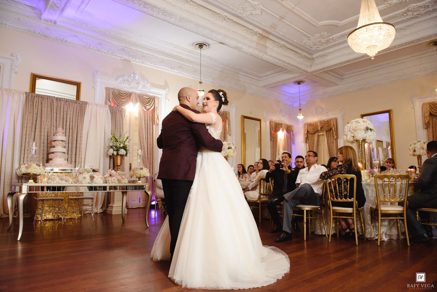 Bride and groom share their first dance in an elegant wedding reception hall with gold chairs and a tiered cake.