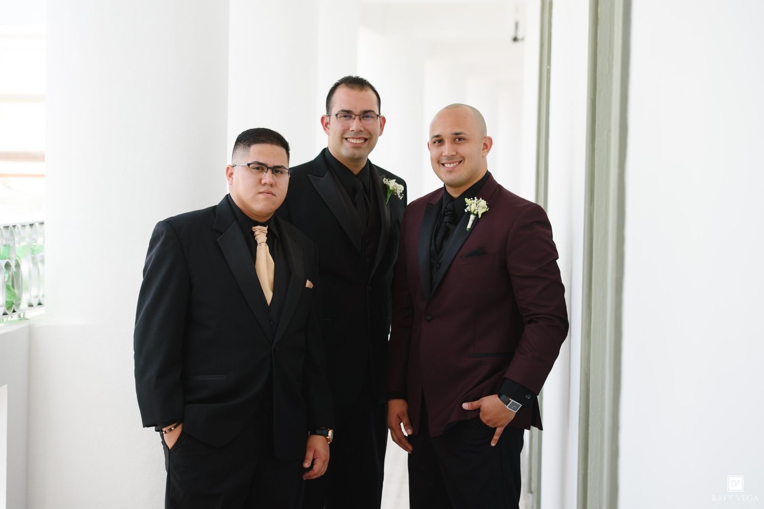Three well-dressed men in suits with boutonnieres posing together at a formal wedding event.