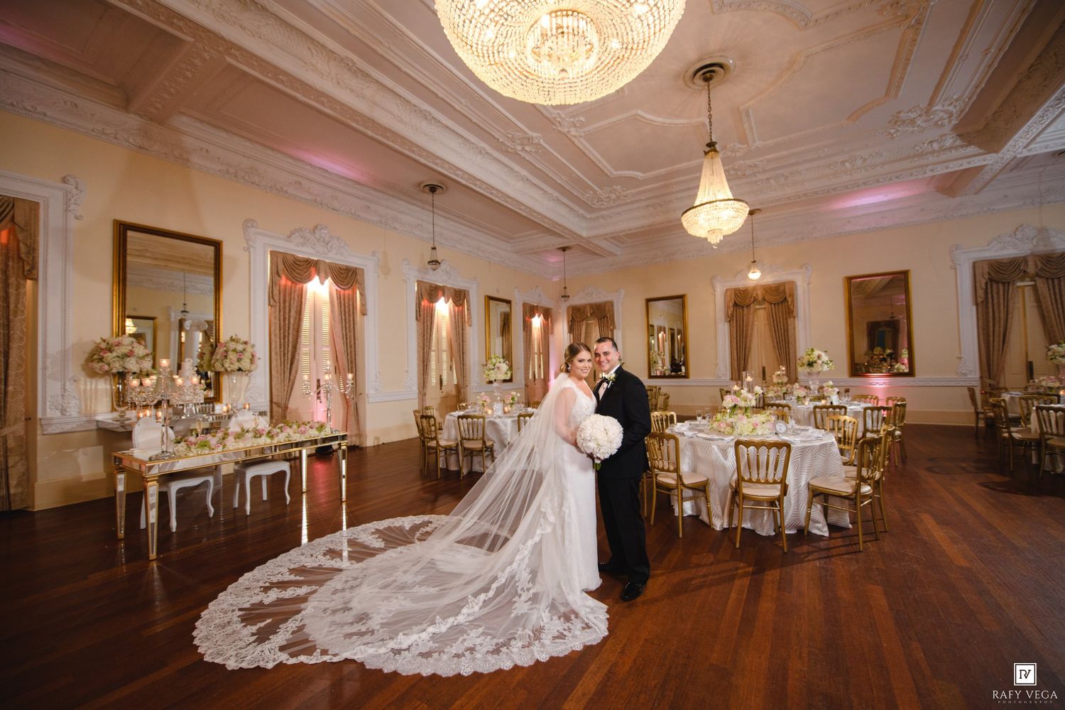 Bride and groom pose together in an elegant ballroom with chandeliers, gold chairs, and floral centerpieces on their wedding day.