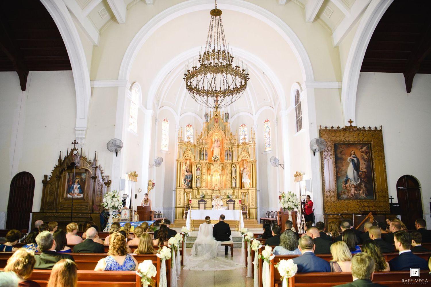 Elegant wedding ceremony inside a historic white church with ornate golden altar, chandelier, and guests seated in wooden pews.