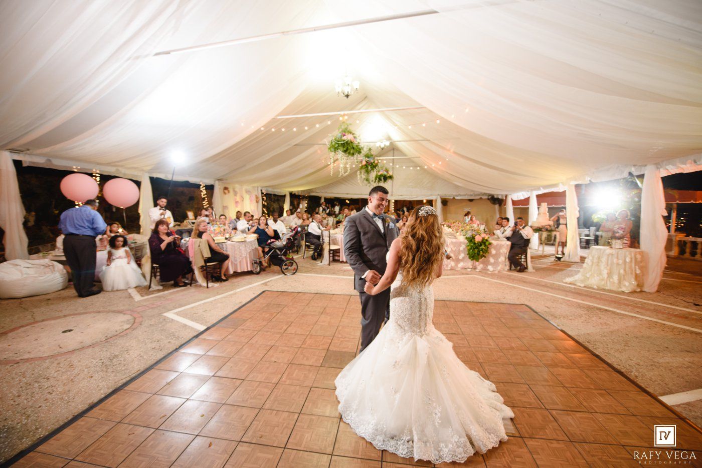 Bride and groom share their first dance on a tile floor beneath a draped white tent with guests watching at wedding reception.