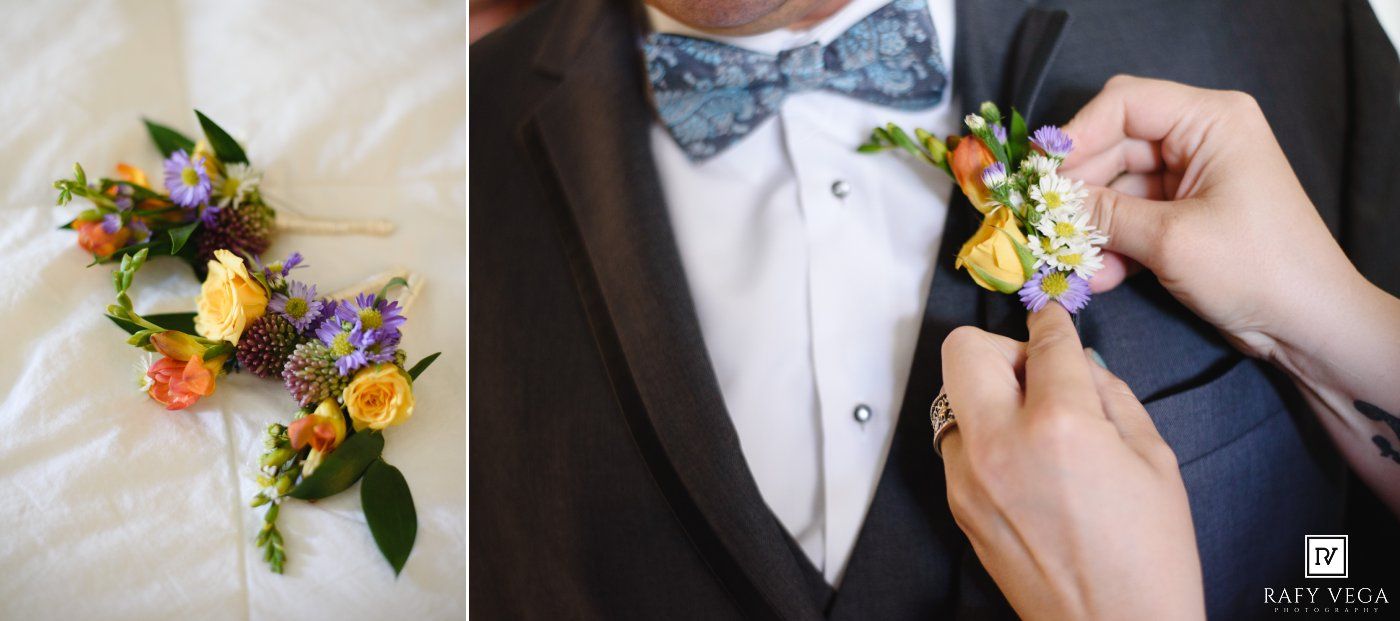 Groom in navy tuxedo having yellow and purple boutonniere pinned, beside a floral wrist corsage on white fabric.
