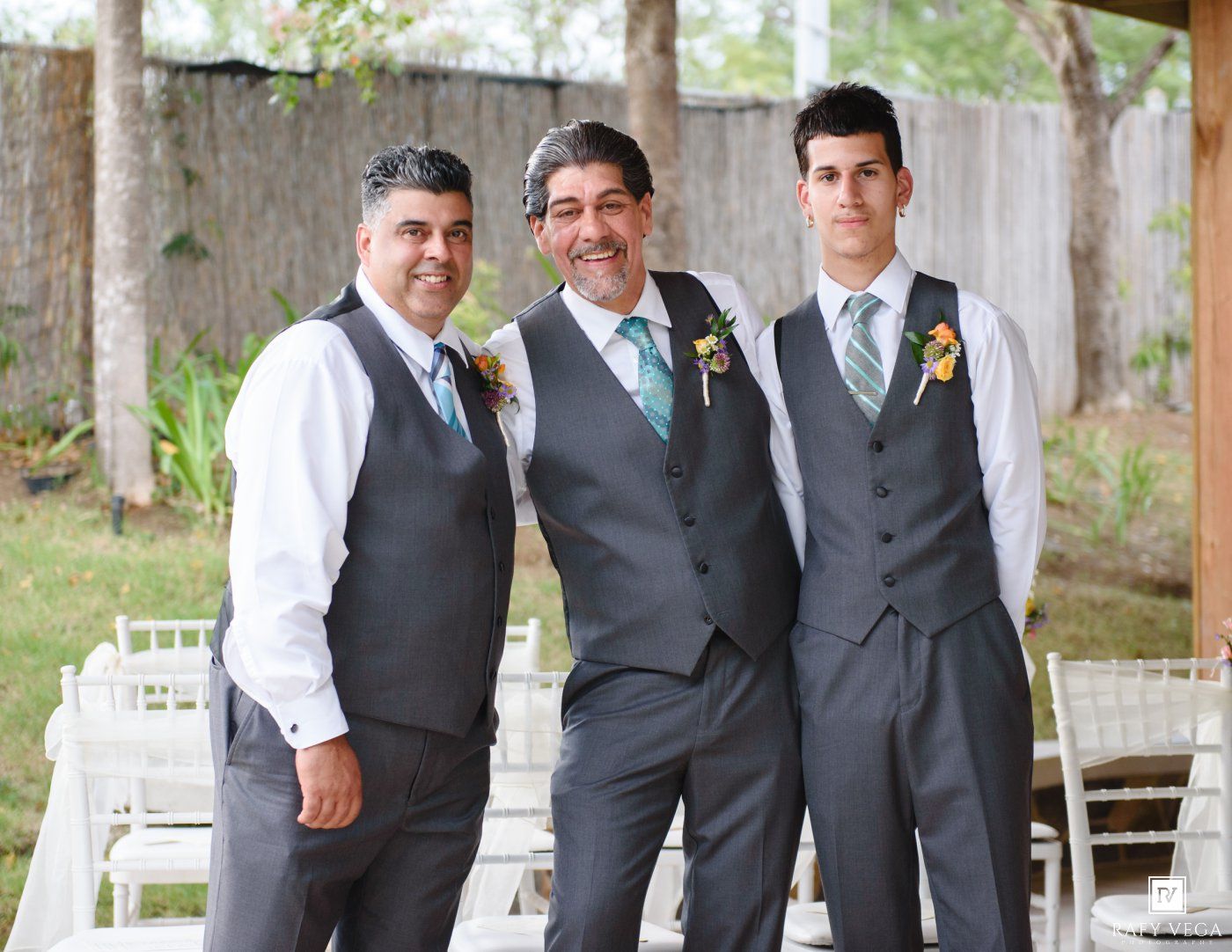 Three groomsmen in gray vests and teal ties smile together outdoors at a wedding venue.
