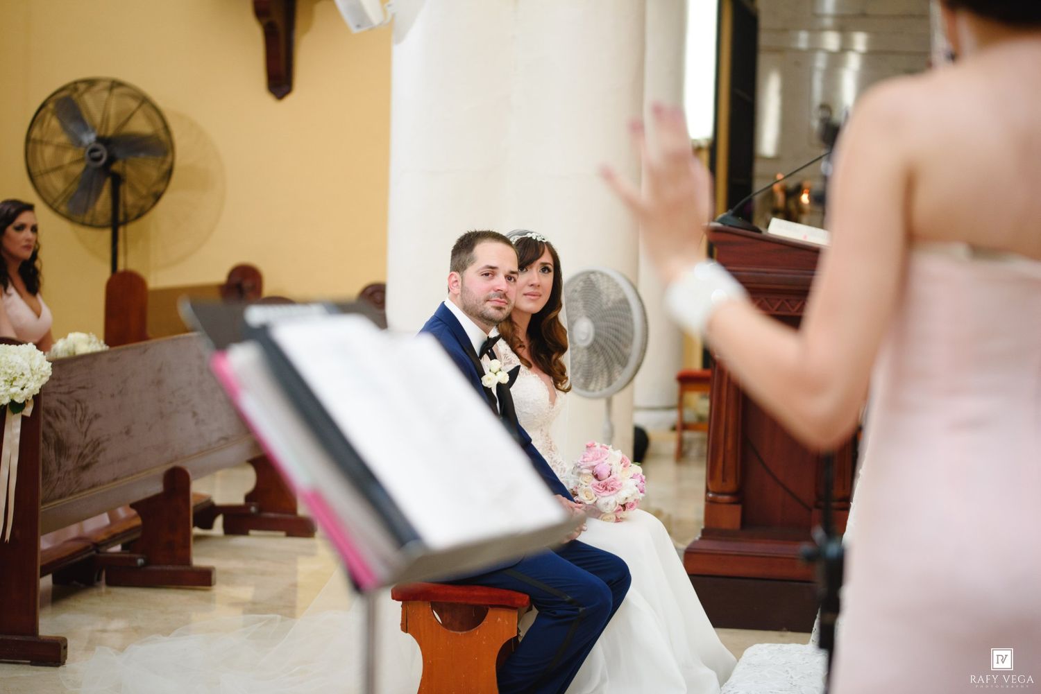 Bride and groom sitting together during church wedding ceremony while a bridesmaid speaks at the podium.