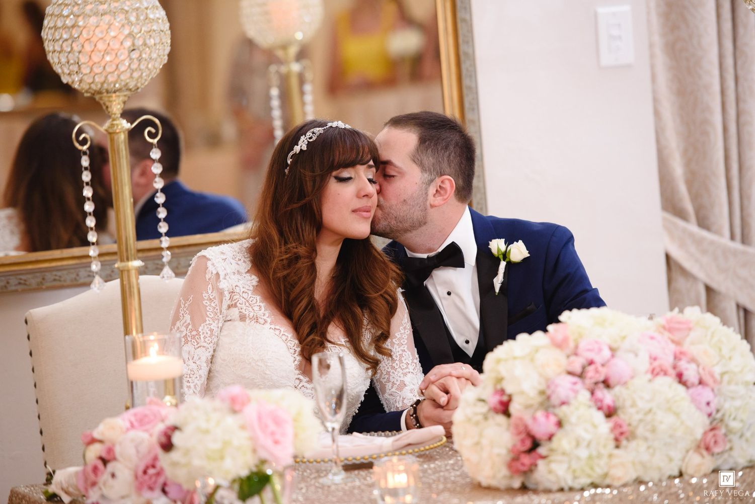 Groom kissing bride at wedding reception sweetheart table with pink floral arrangements and crystal candelabra.