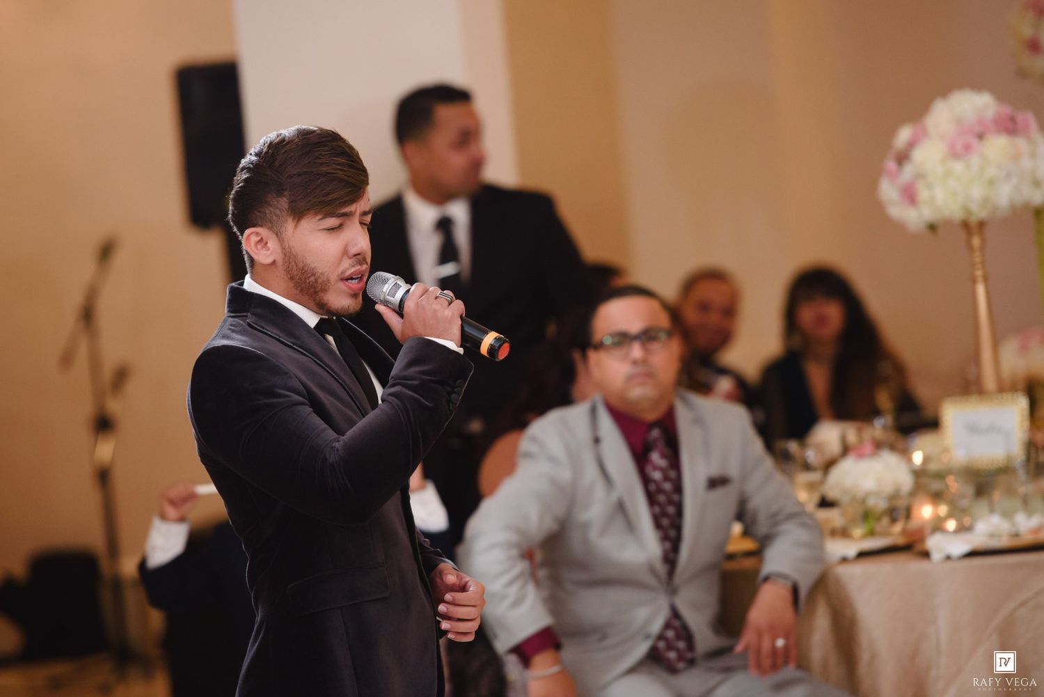 Man in black suit speaking into microphone at elegant wedding reception with floral centerpieces.