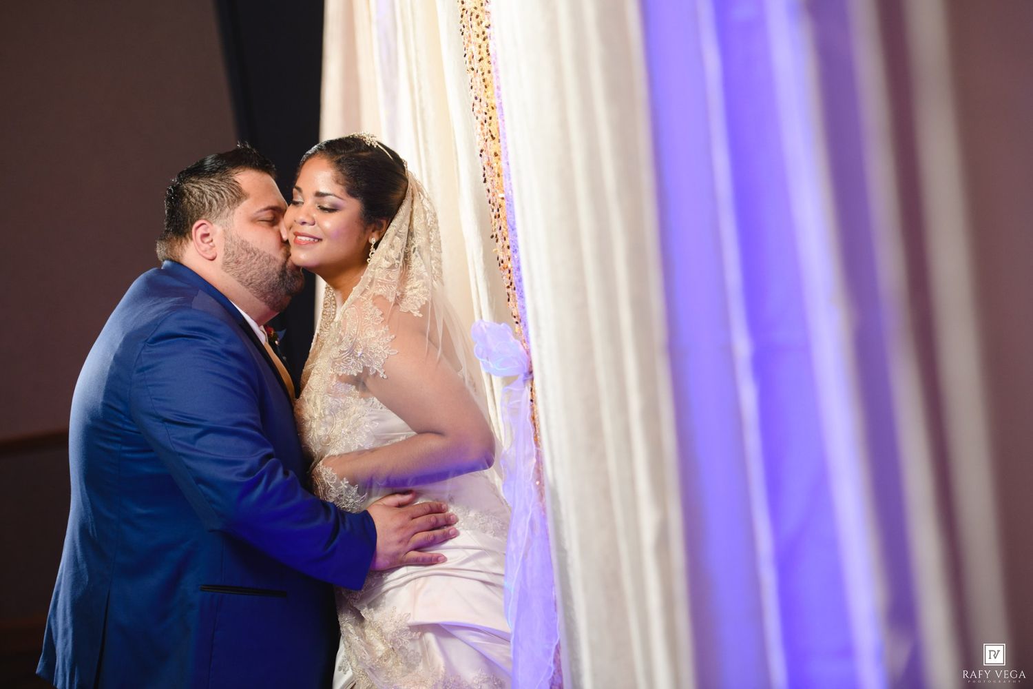 A groom in a blue suit kisses his bride in a white lace gown and veil at a wedding ceremony.