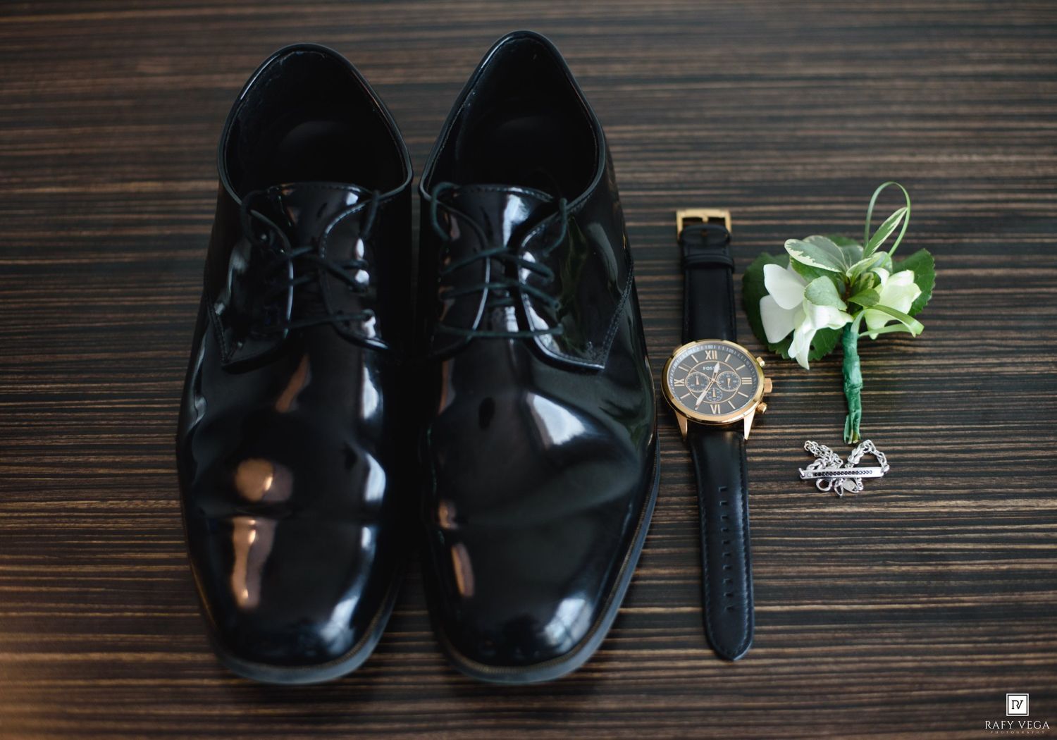 Black patent leather dress shoes beside a gold chronograph watch, boutonniere, and cufflinks on wooden surface.
