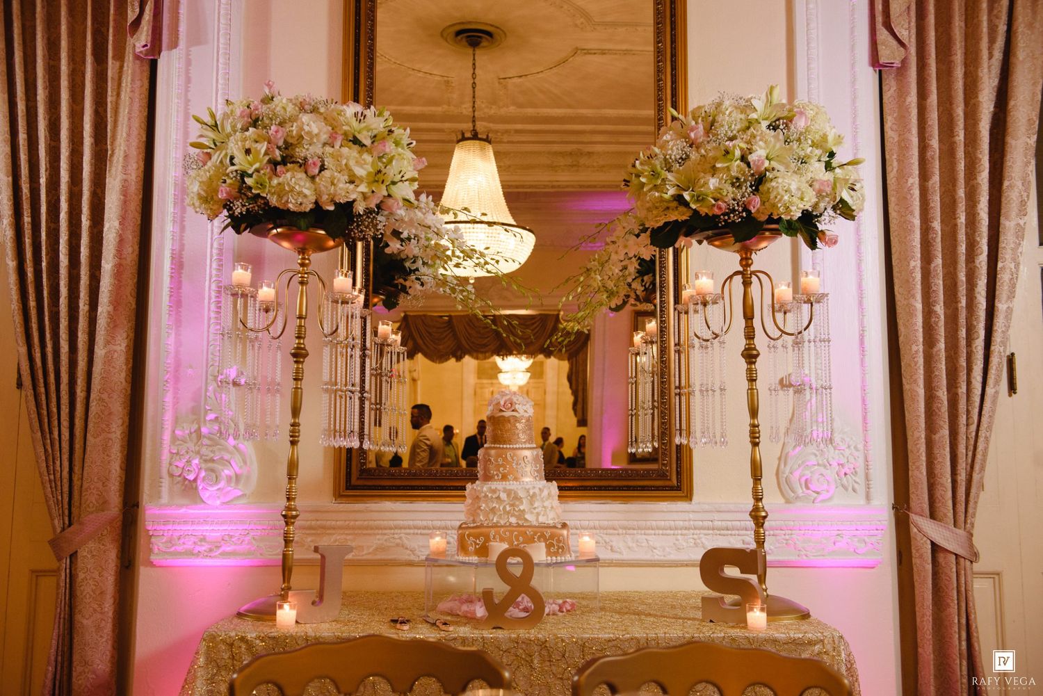 Elegant wedding cake display with tall gold candelabras, white floral arrangements, and pink uplighting in a luxurious ballroom.