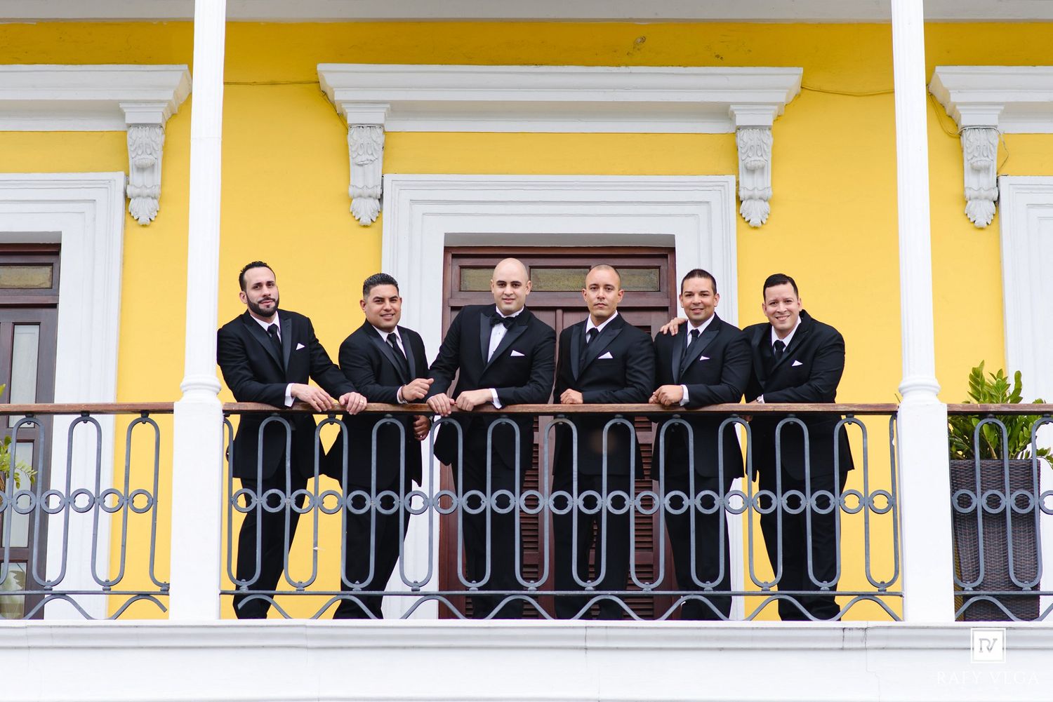 Six groomsmen in black suits pose on a decorative balcony of a yellow building for a wedding photo.