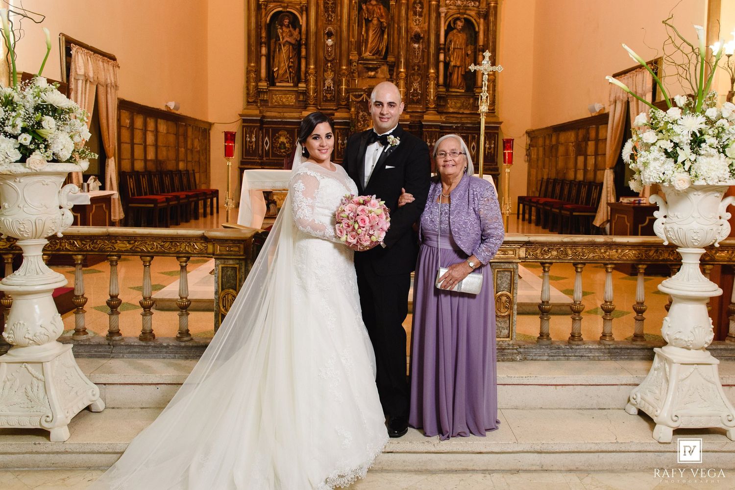 Bride in white gown and groom in tuxedo pose with older woman in lavender dress inside ornate church.