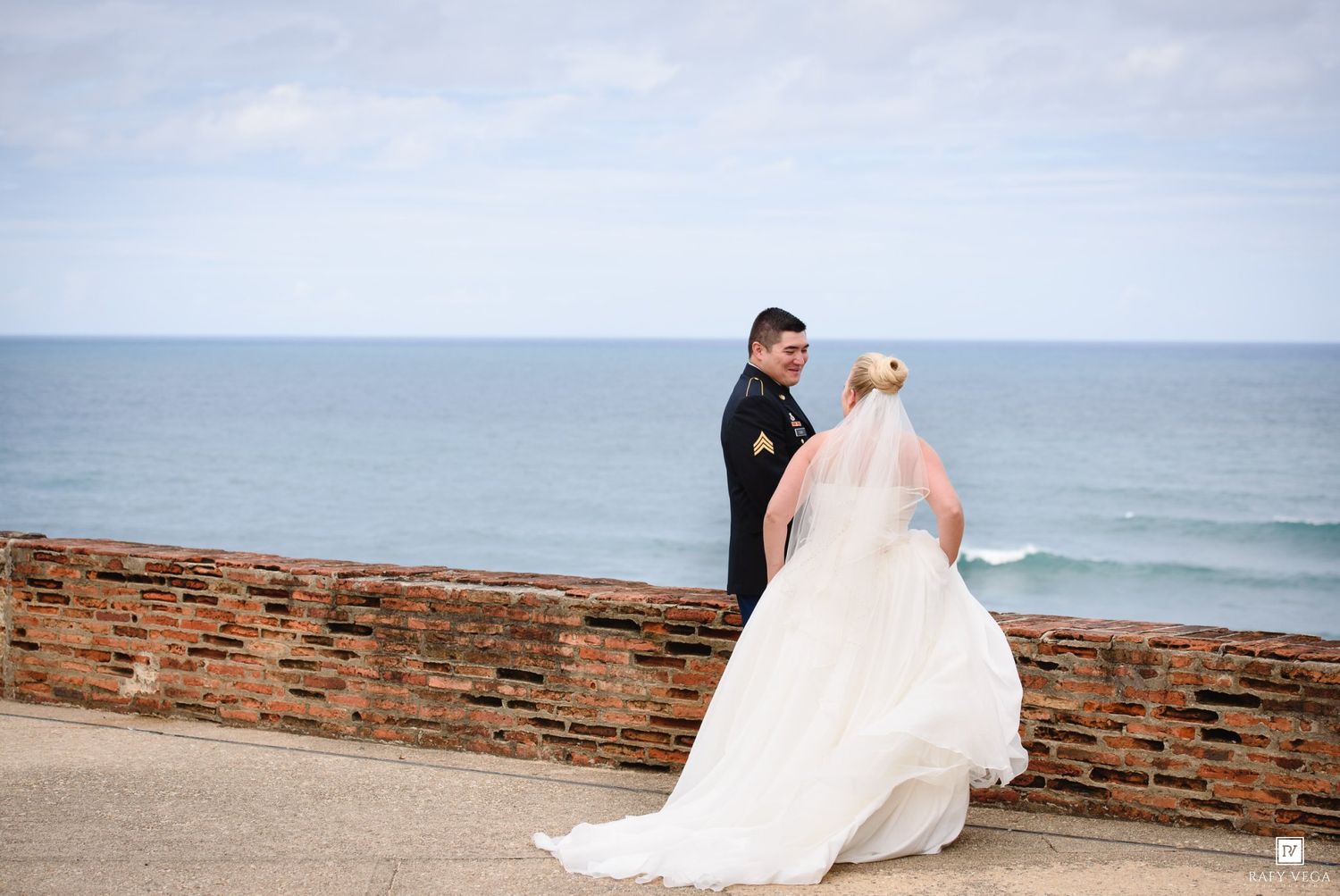 Newlyweds share a romantic moment by a seaside brick wall, bride in flowing white gown.